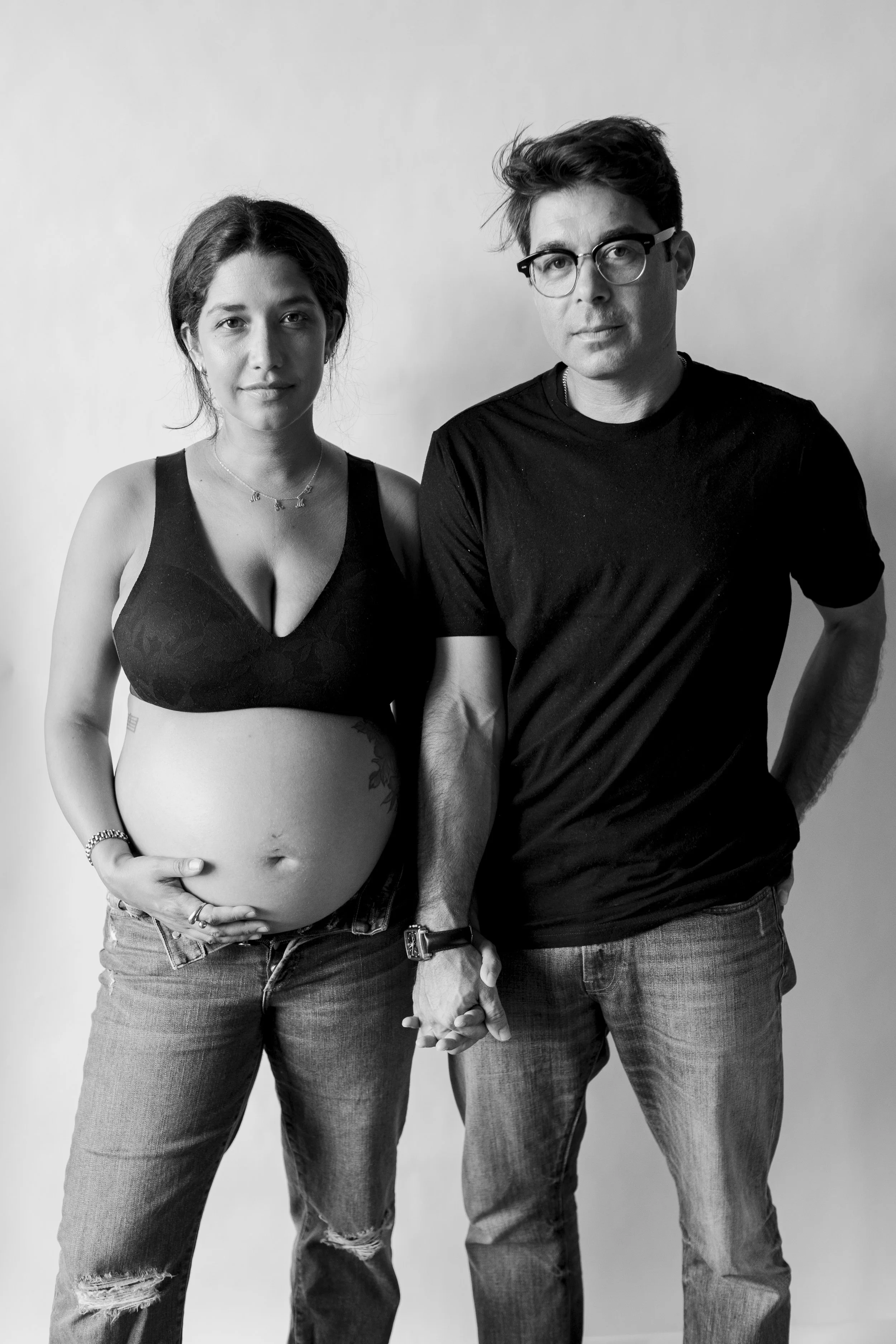 A black and white photo of a pregnant woman holding her belly, standing beside a man. They are holding hands and facing forward, against a plain background.