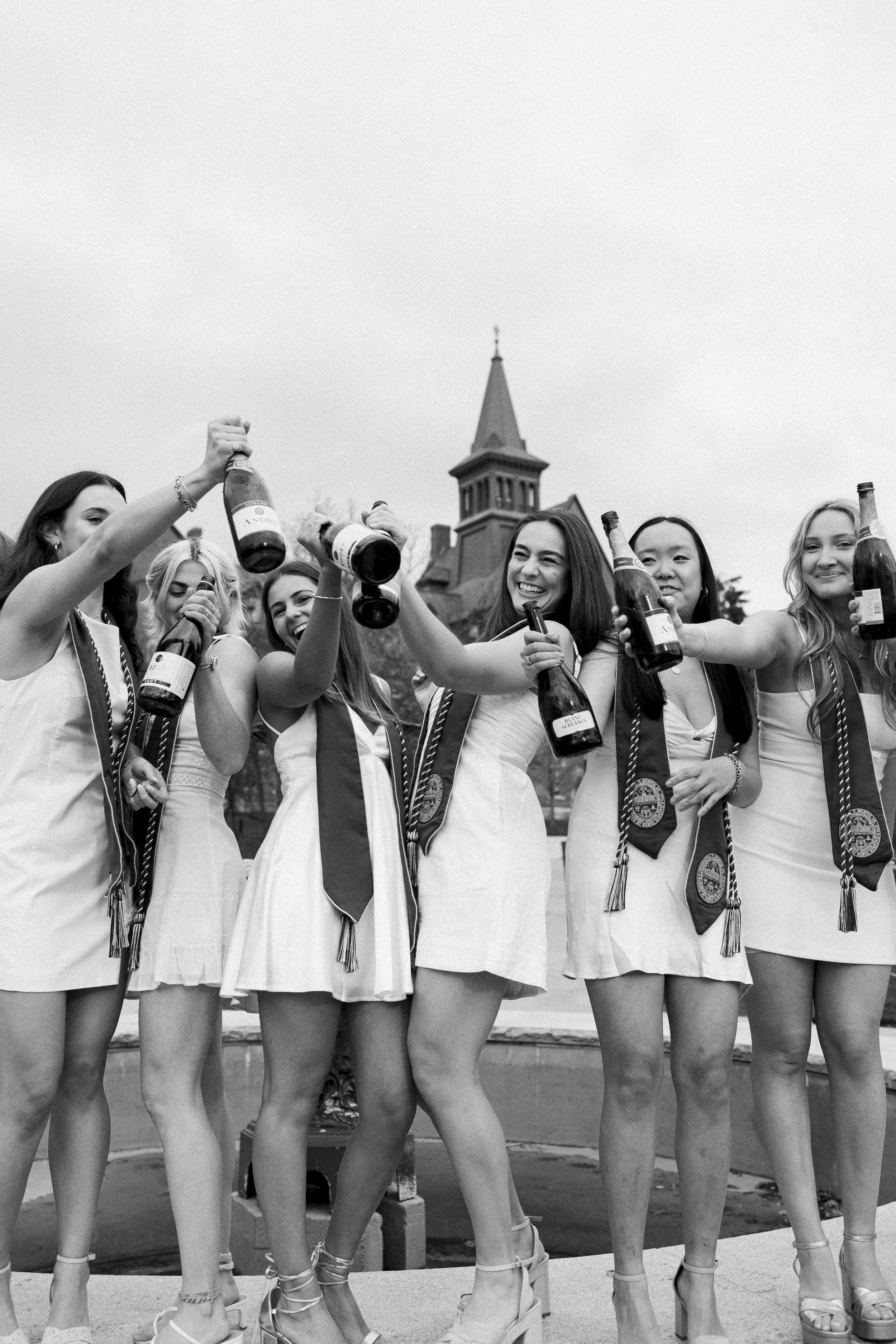 A group of young women in white dresses celebrating together outdoors, holding bottles of champagne and toasting.