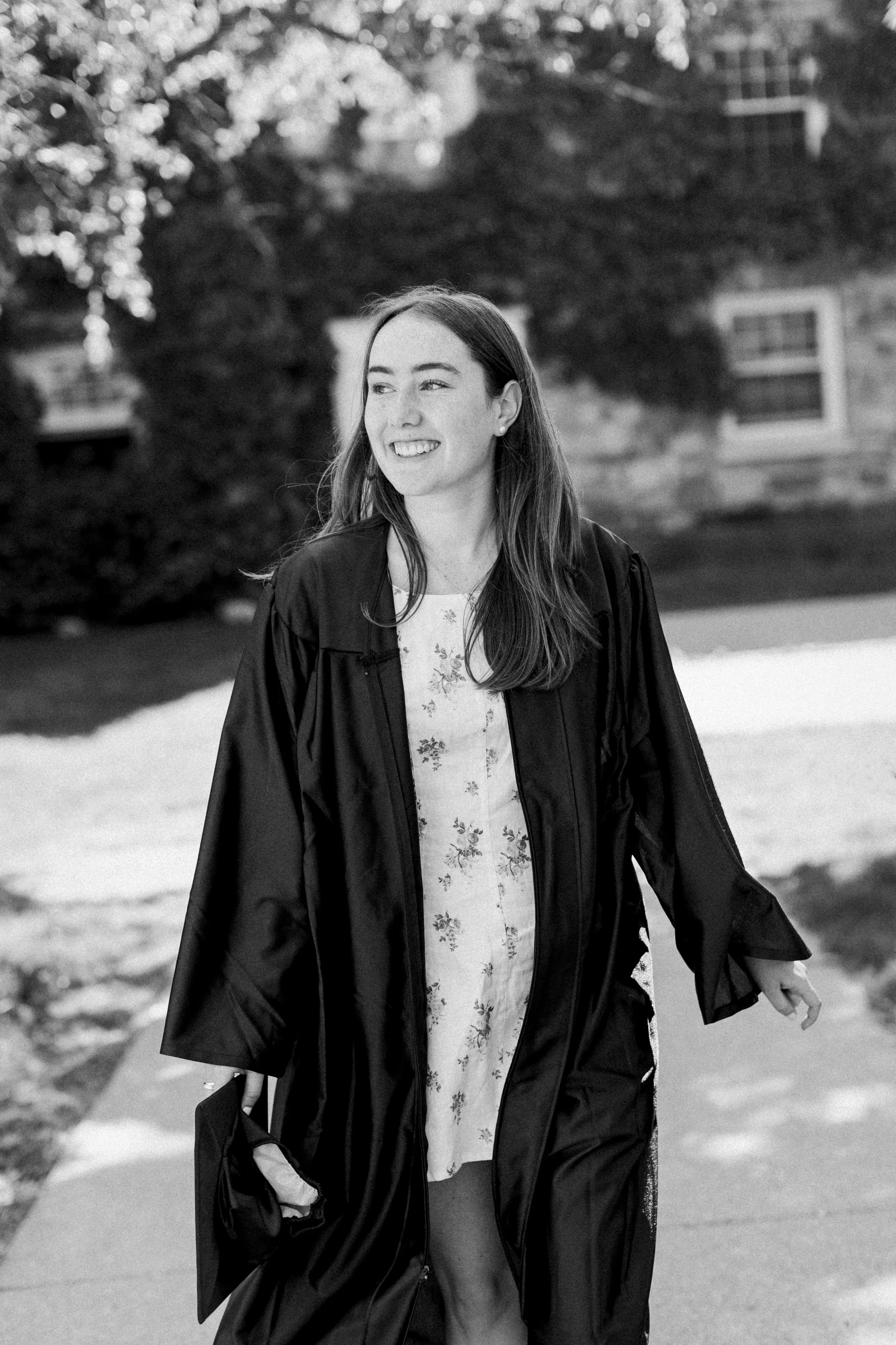 Black and white photo of a young woman in a graduation gown and cap, smiling and walking outdoors.