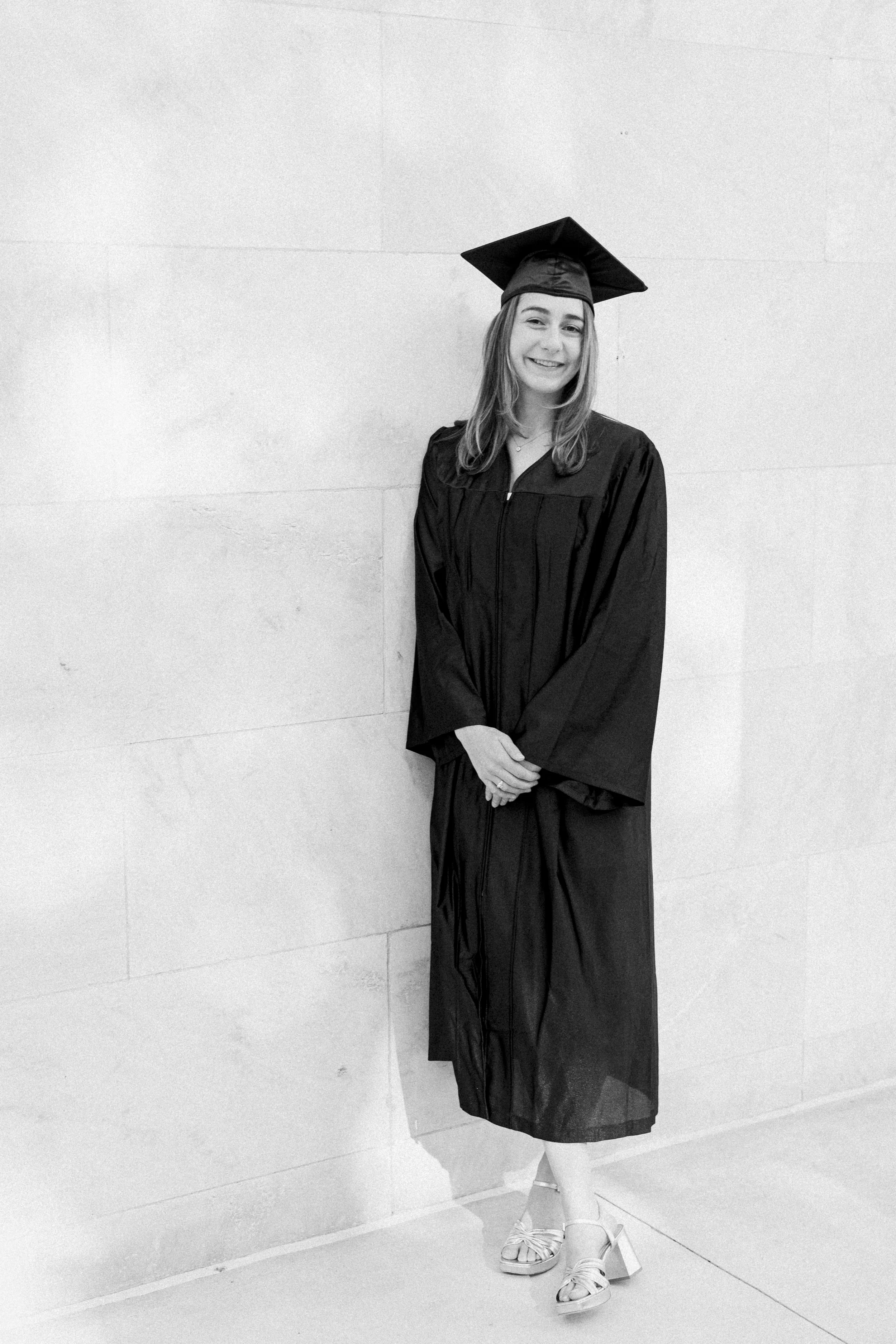 A young woman in a graduation gown and cap standing against a wall, smiling at the camera.