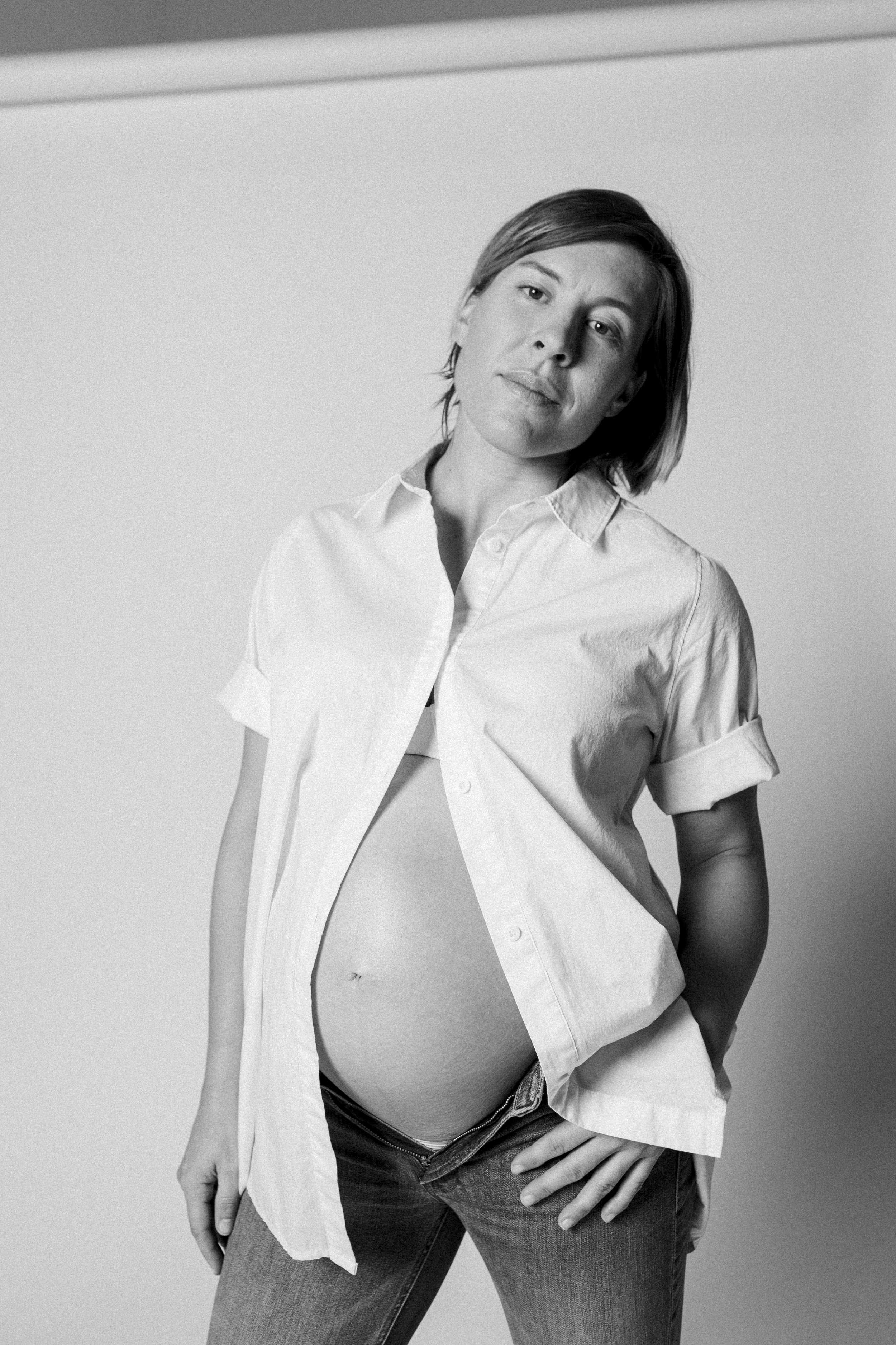 Black and white photograph of a woman with short hair, wearing an unbuttoned white shirt and jeans, standing against a plain background.
