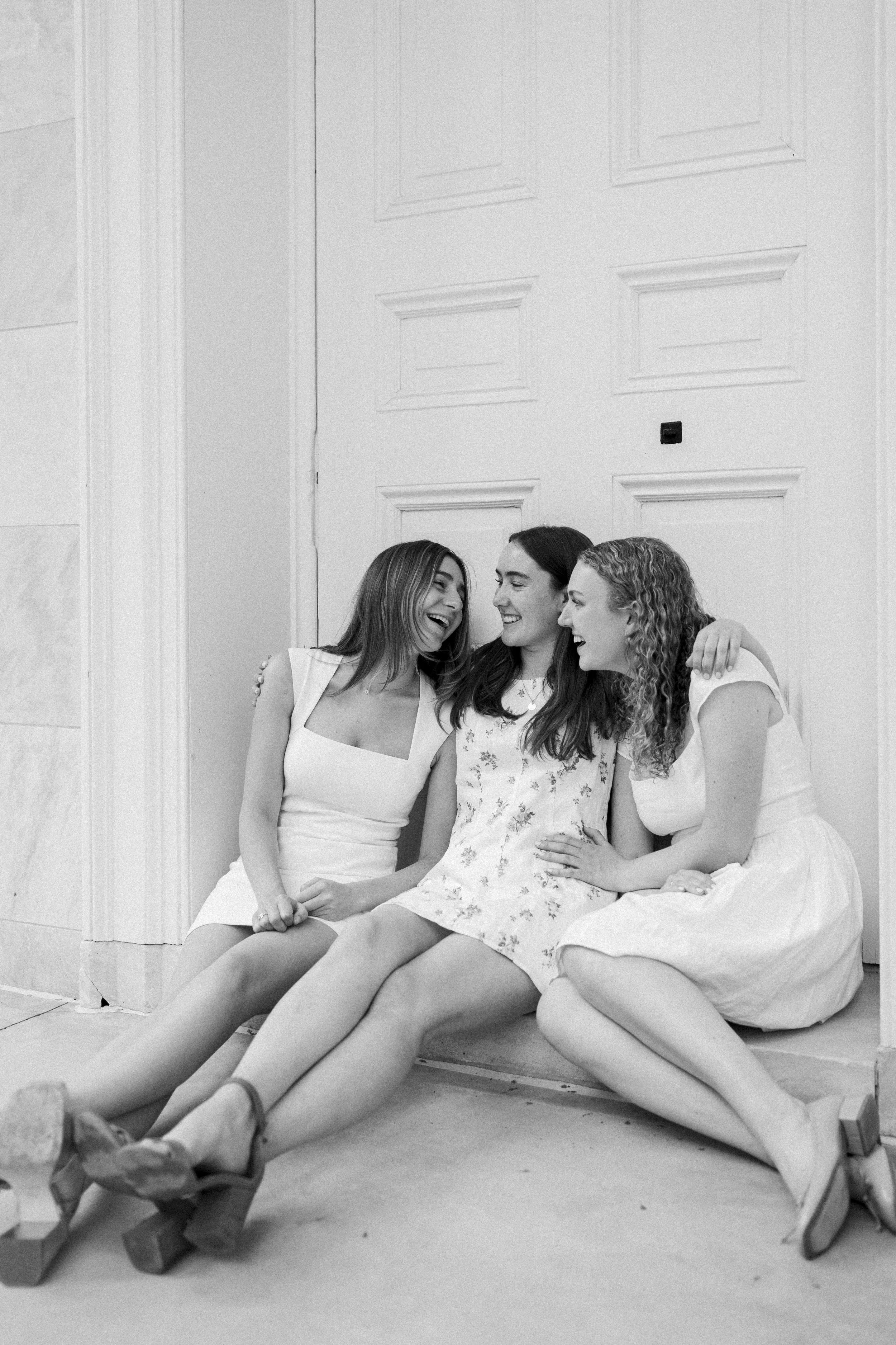 Three women sitting on a step, laughing and hugging each other, in front of a closed door.