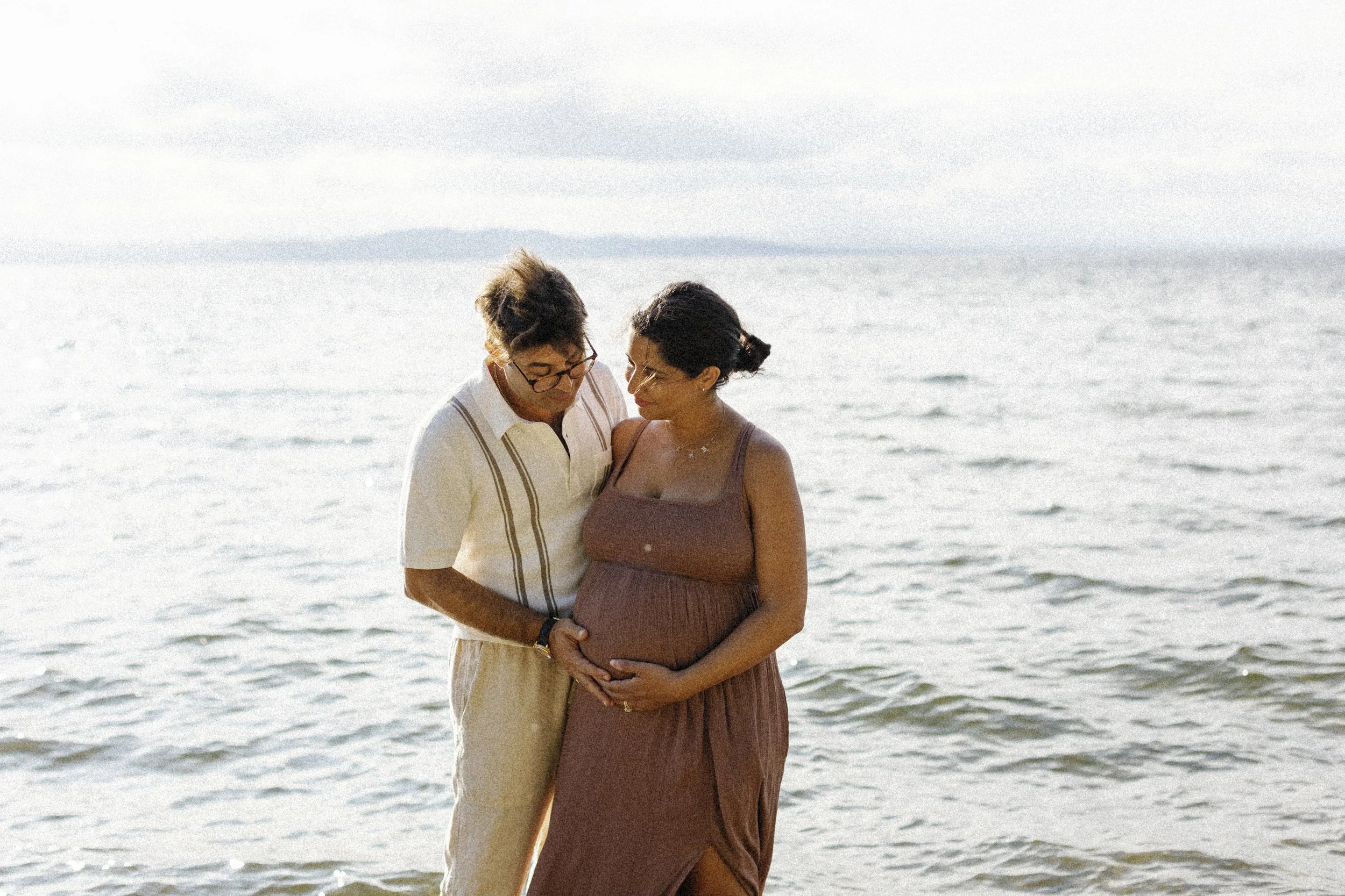A couple standing in the water at the beach, with the man touching the pregnant woman's belly, both looking down lovingly.