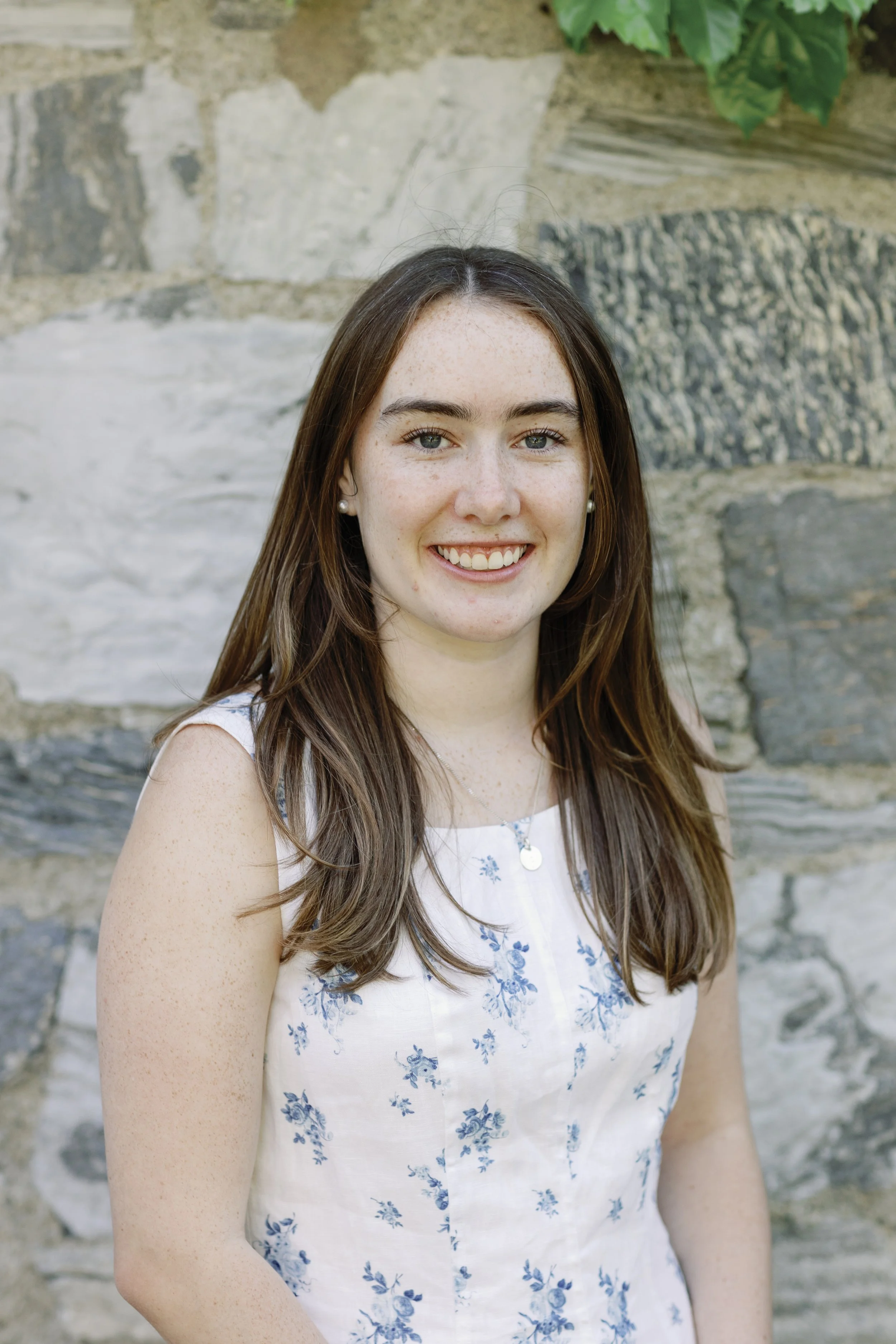 Smiling young woman with long brown hair, wearing a white sleeveless dress with blue floral patterns and pearl earrings, standing outdoors in front of a stone wall with green foliage.