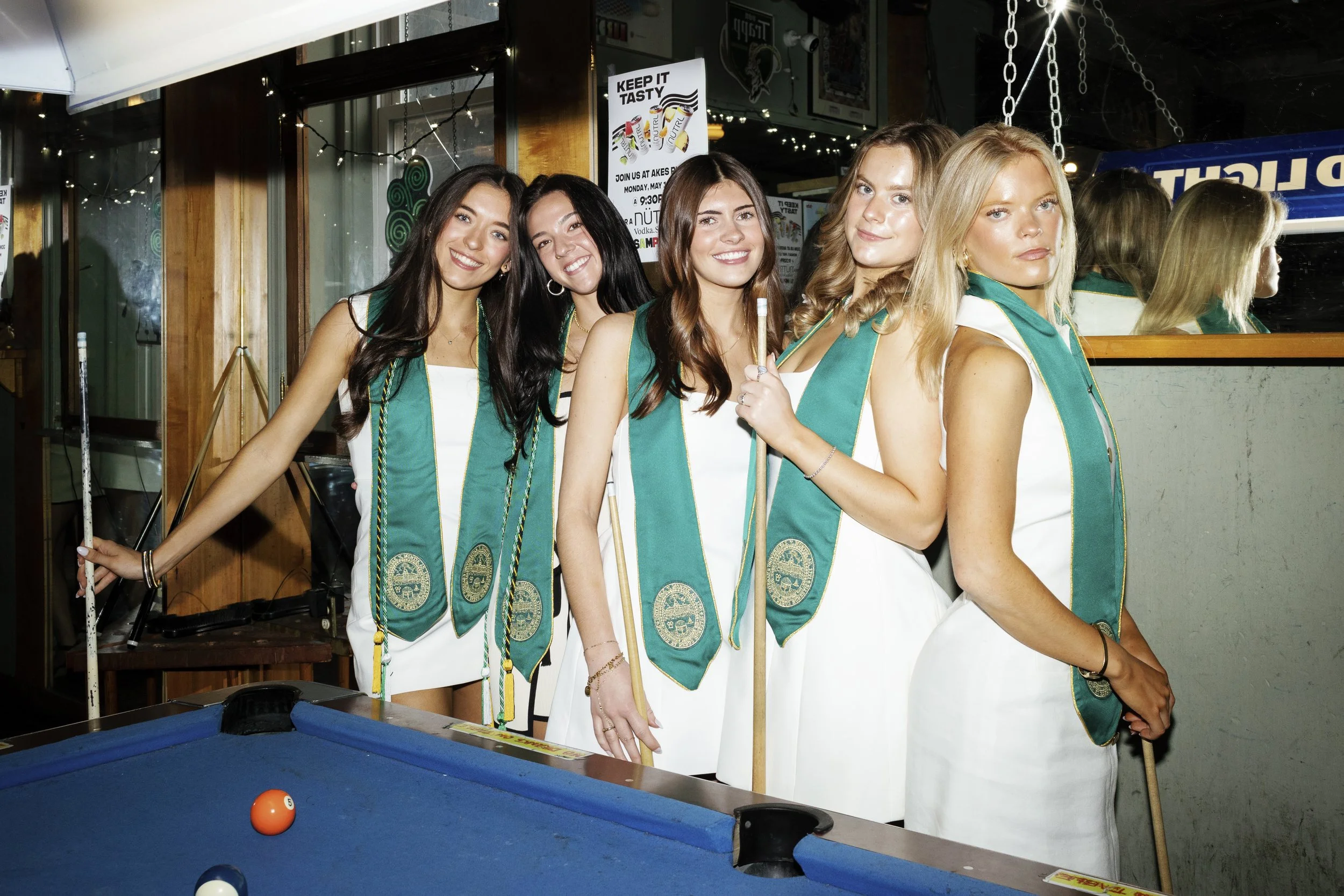 Five young women wearing white dresses and green sashes standing around a pool table in a bar, smiling at the camera.