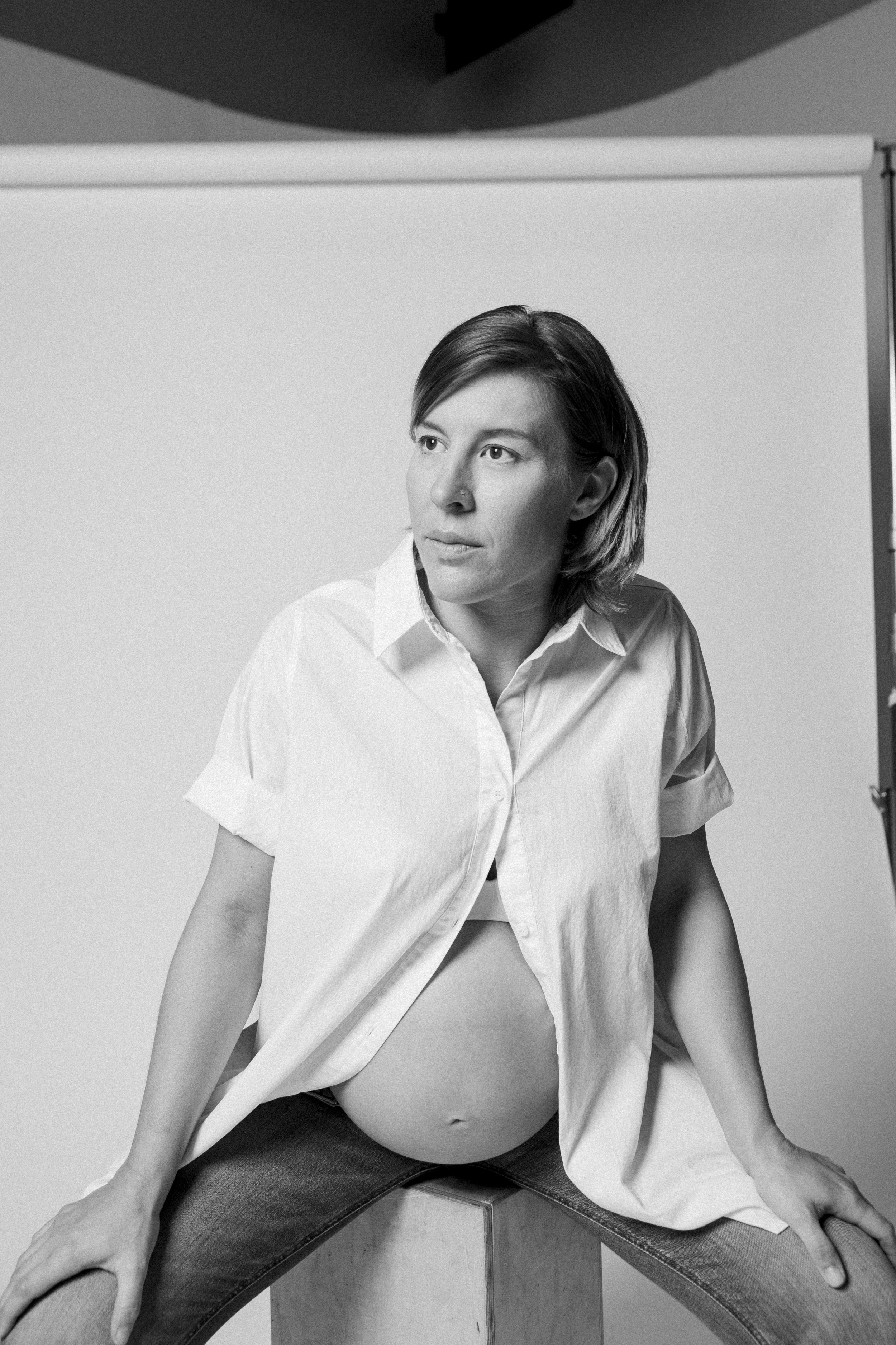 Black and white photo of a pregnant woman sitting on a cube, wearing a partially unbuttoned white shirt, with a plain backdrop.