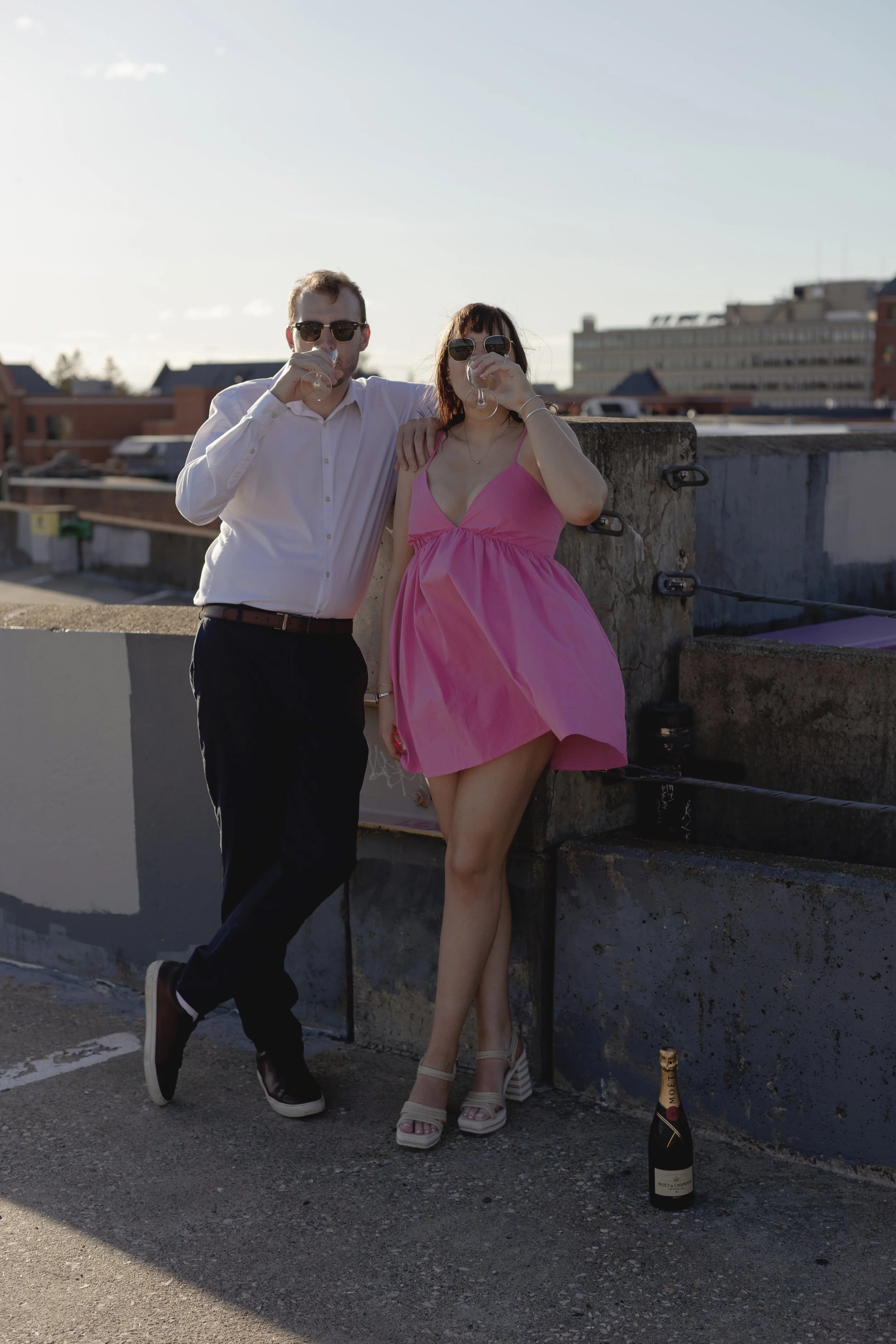 A man and woman on a rooftop during sunset, drinking champagne. The man is wearing sunglasses, a white shirt, and dark pants, with his arm around the woman. The woman is dressed in a pink dress and high heels, holding a glass of wine. A bottle of champagne is placed on the ground nearby.