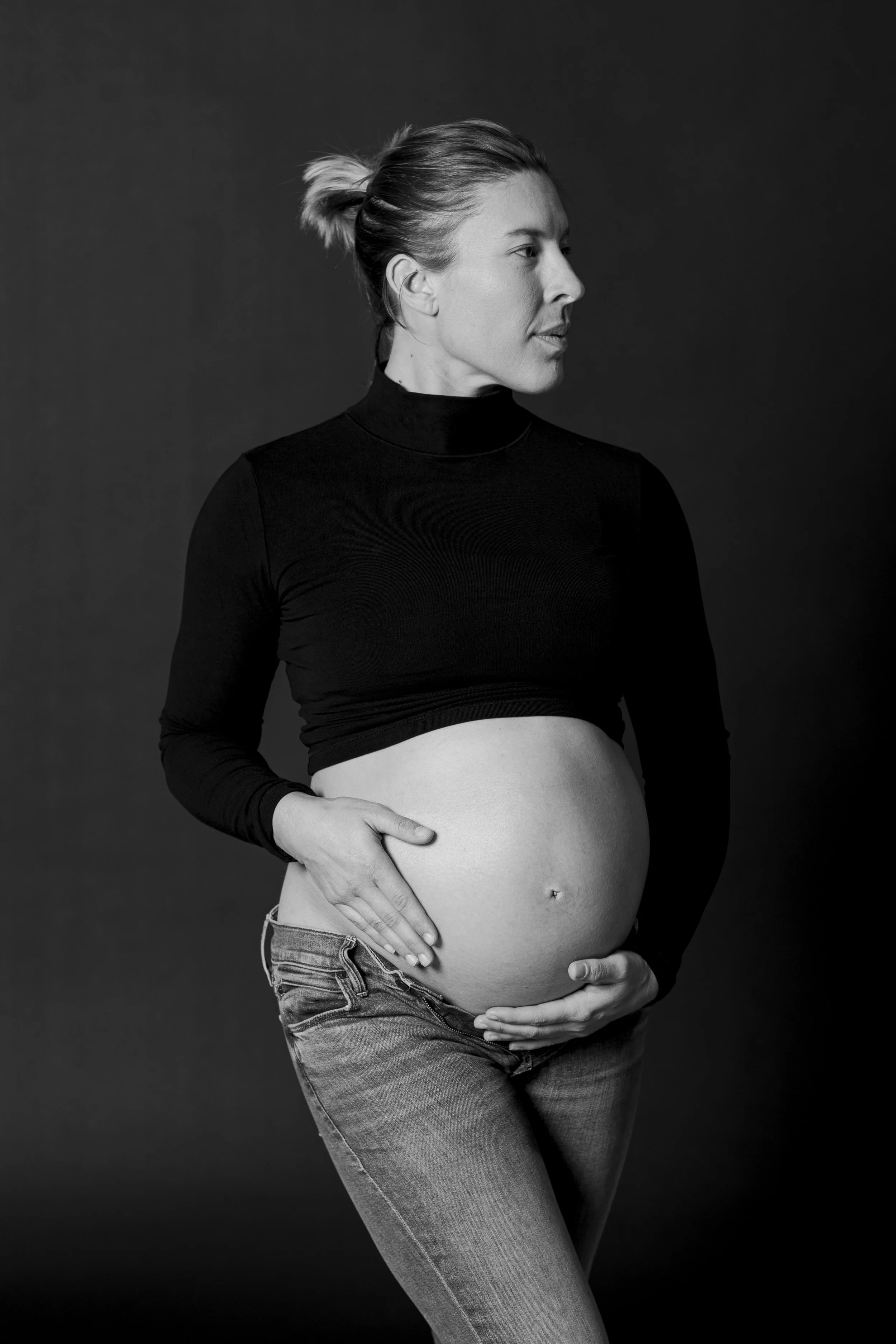 Black and white portrait of a pregnant woman with her hand on her belly, wearing a black long-sleeve top and jeans, looking to the side against a plain background.