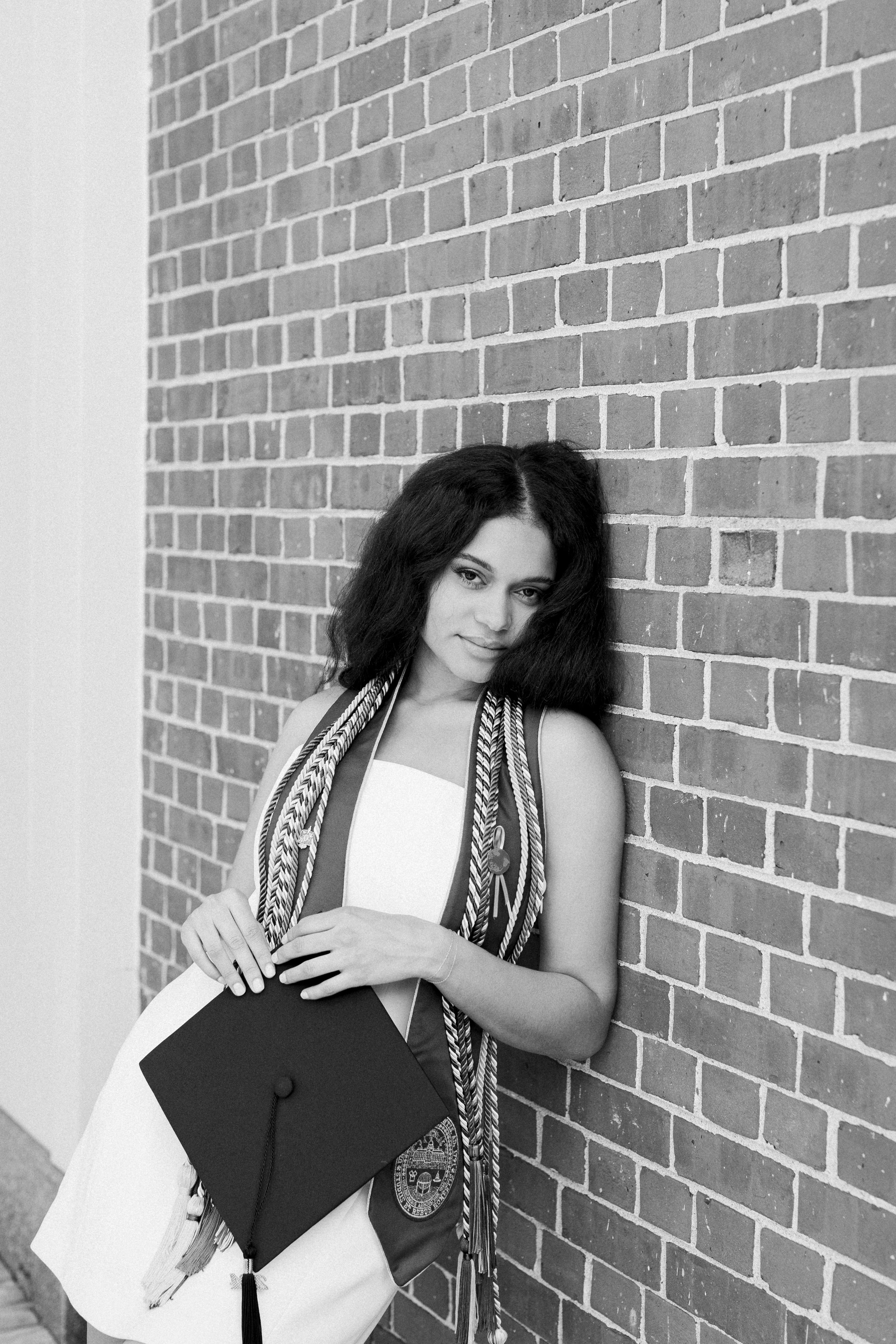 A young woman in graduation attire holding a cap, leaning against a brick wall, smiling softly.