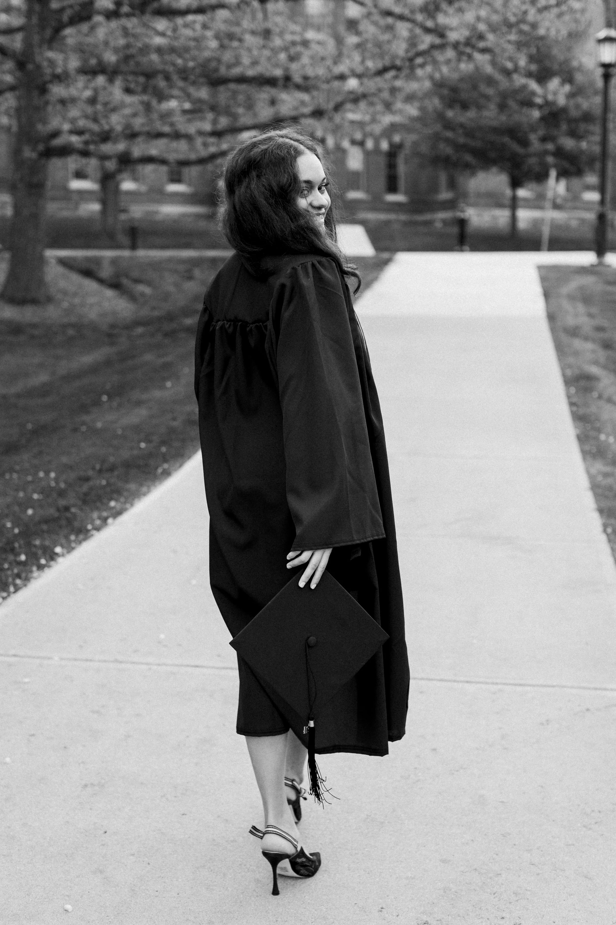 A woman in a graduation gown and cap walking on a campus sidewalk, holding her cap in her hand, with trees and buildings in the background.