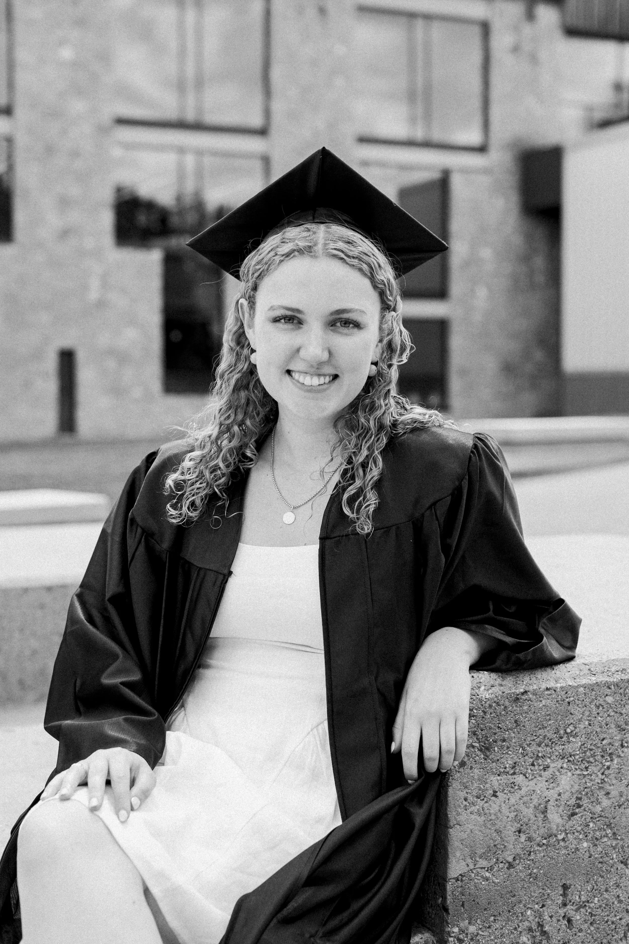 A young woman in graduation cap and gown sitting on a concrete ledge outdoors, smiling at the camera.