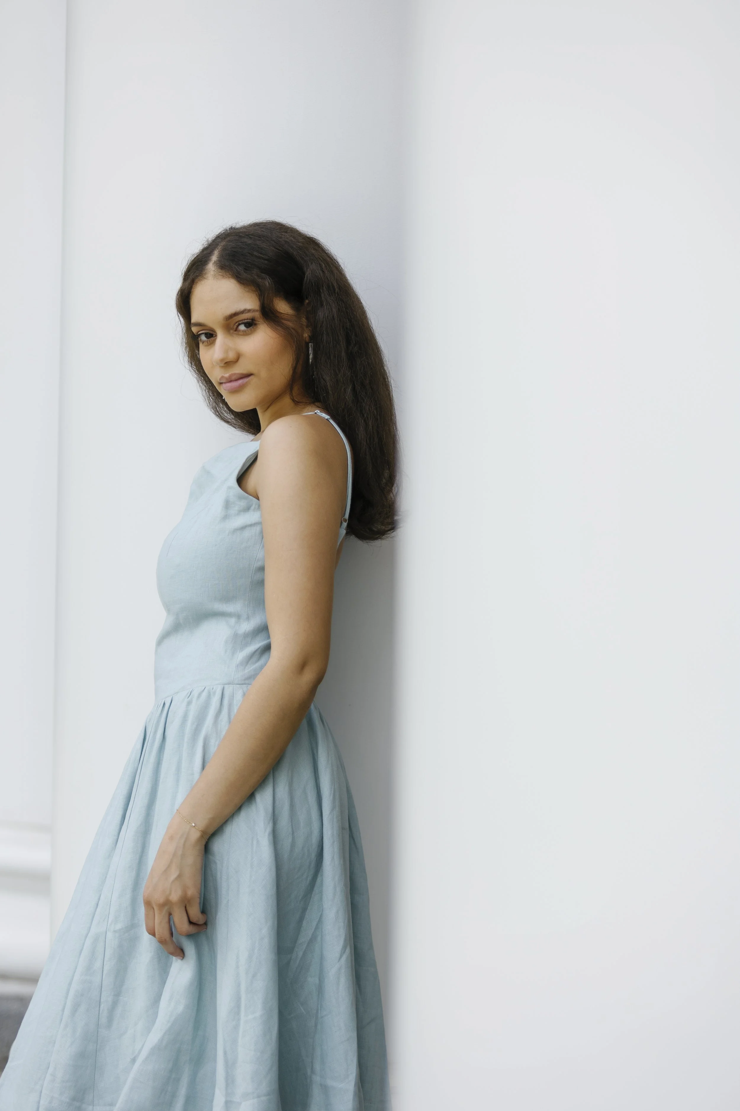 A woman with dark wavy hair wearing a light blue dress, standing with her back against a plain white wall.
