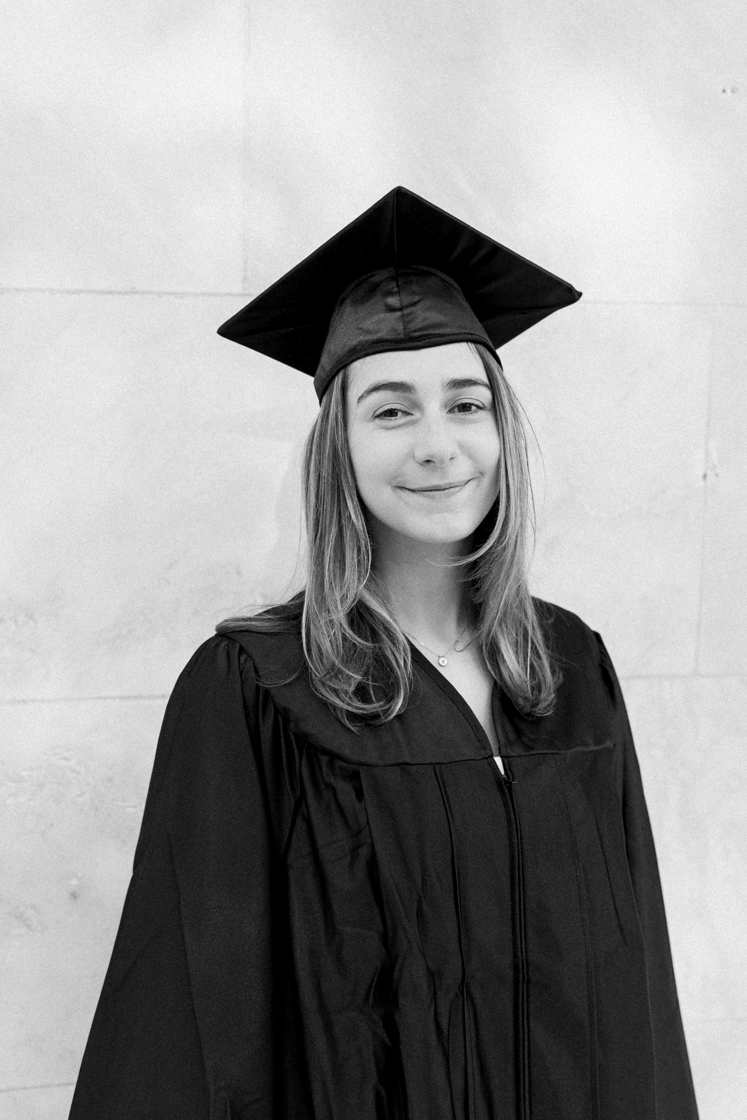Black and white photo of a young woman in a graduation cap and gown, smiling softly, standing against a plain wall.