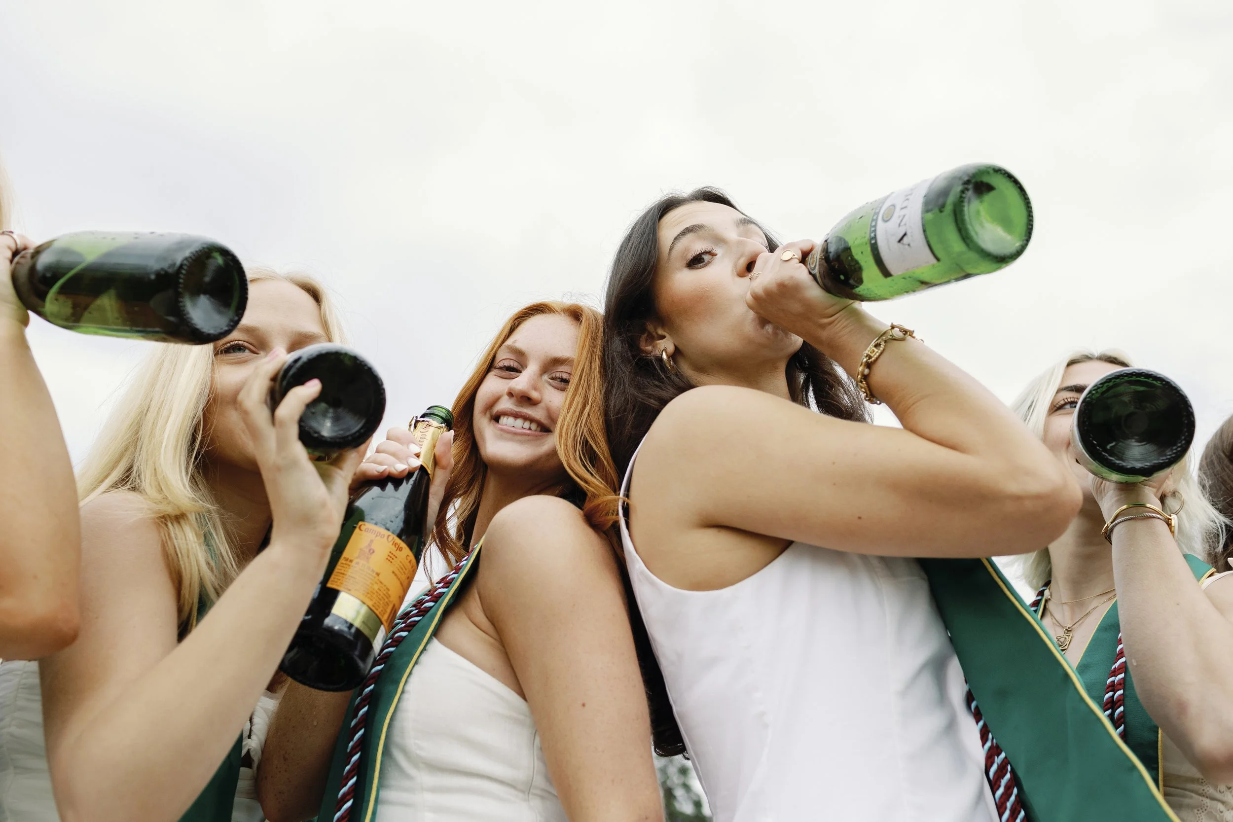 Group of women celebrating outdoors, drinking from bottles, smiling, dressed in casual summer clothes.