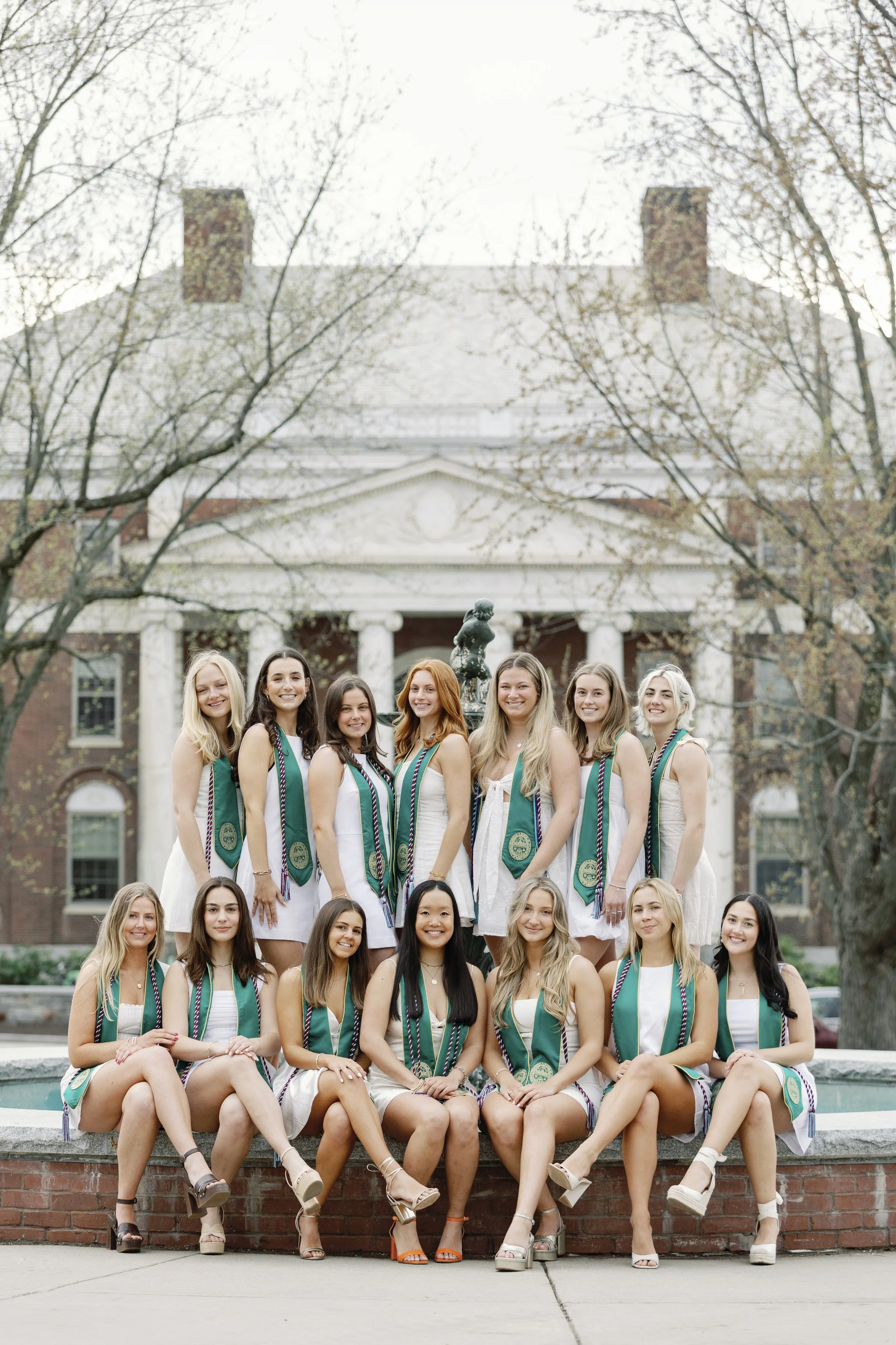 Group of young women wearing white dresses and green graduation stoles posing in front of a historic building with trees in background.