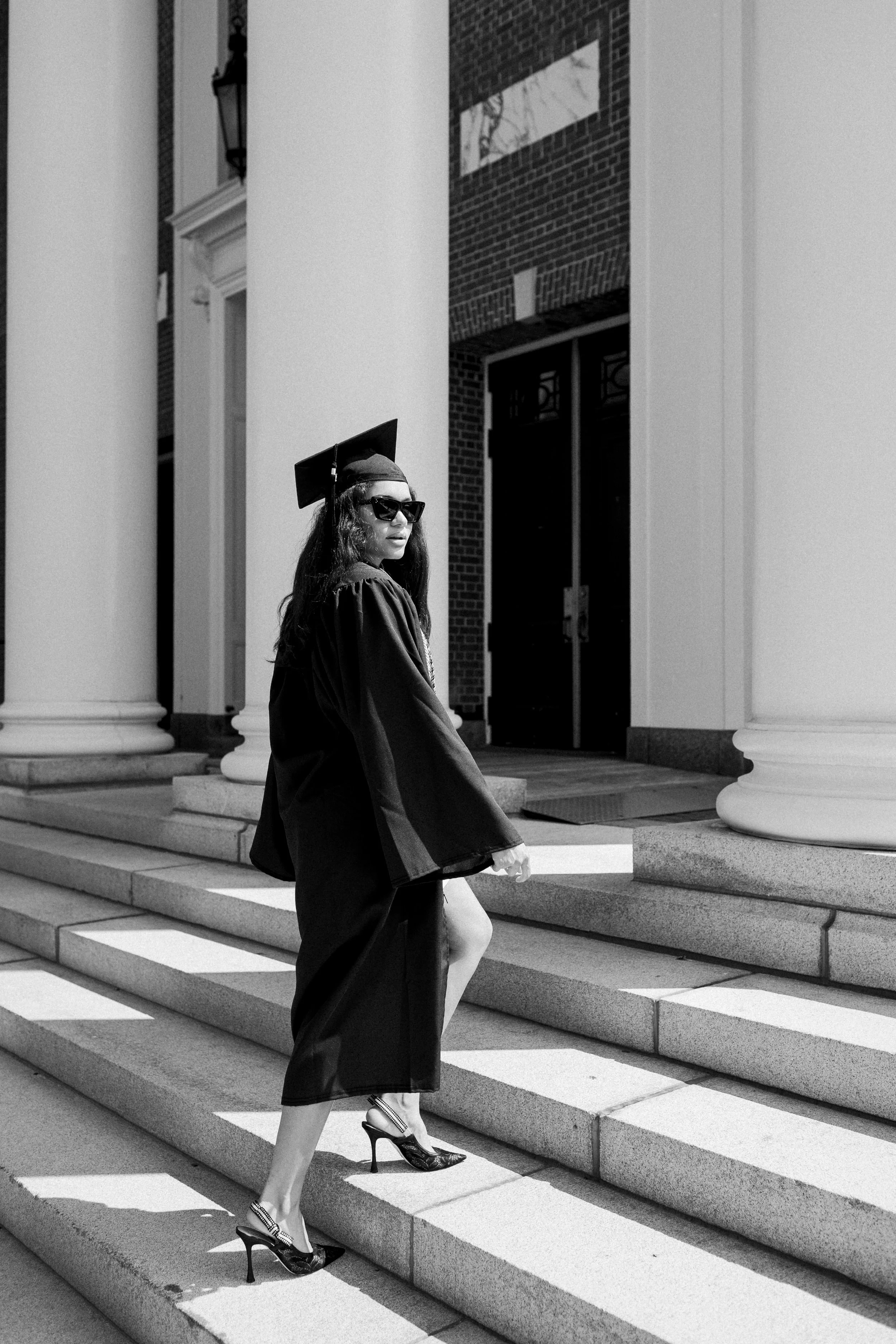 Young woman in graduation cap and gown walking up steps outside a building with large columns.