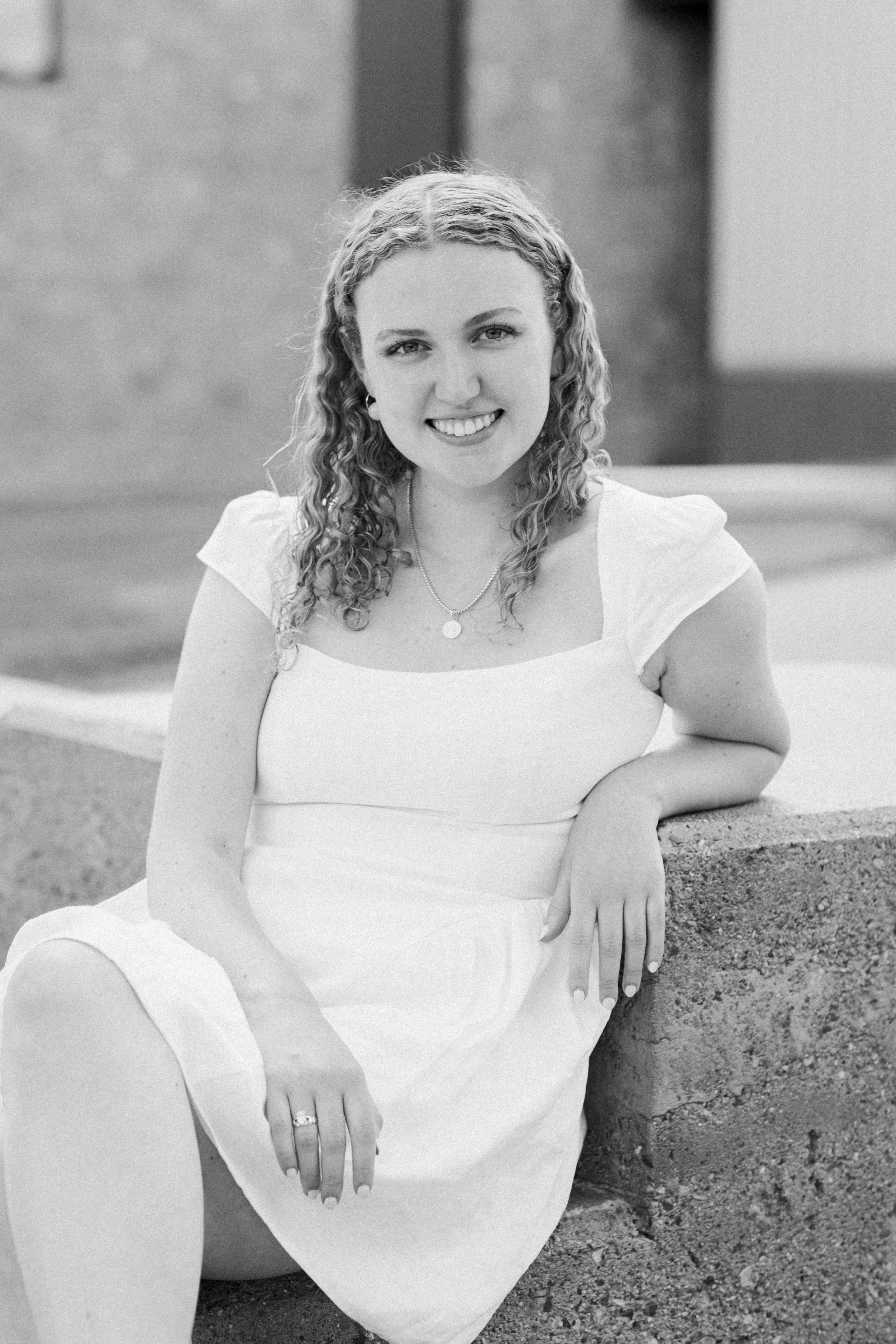 Black and white photo of a young woman sitting on a concrete ledge, smiling, wearing a white dress and a necklace, outdoors in front of a blurred background.