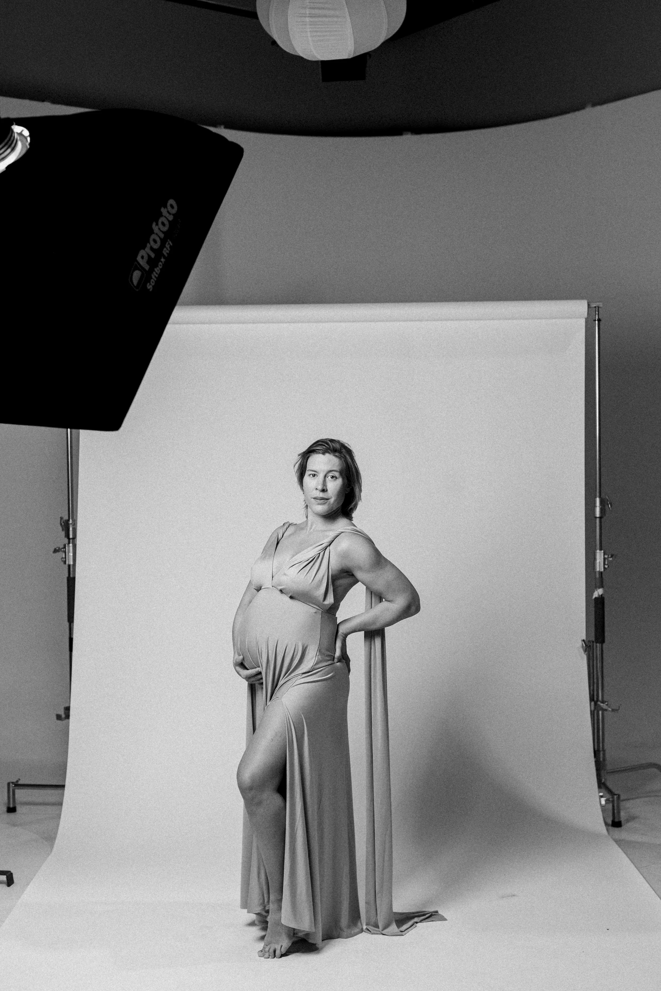 A pregnant woman in a flowing dress posing in a studio with a white backdrop, professional lighting equipment nearby.