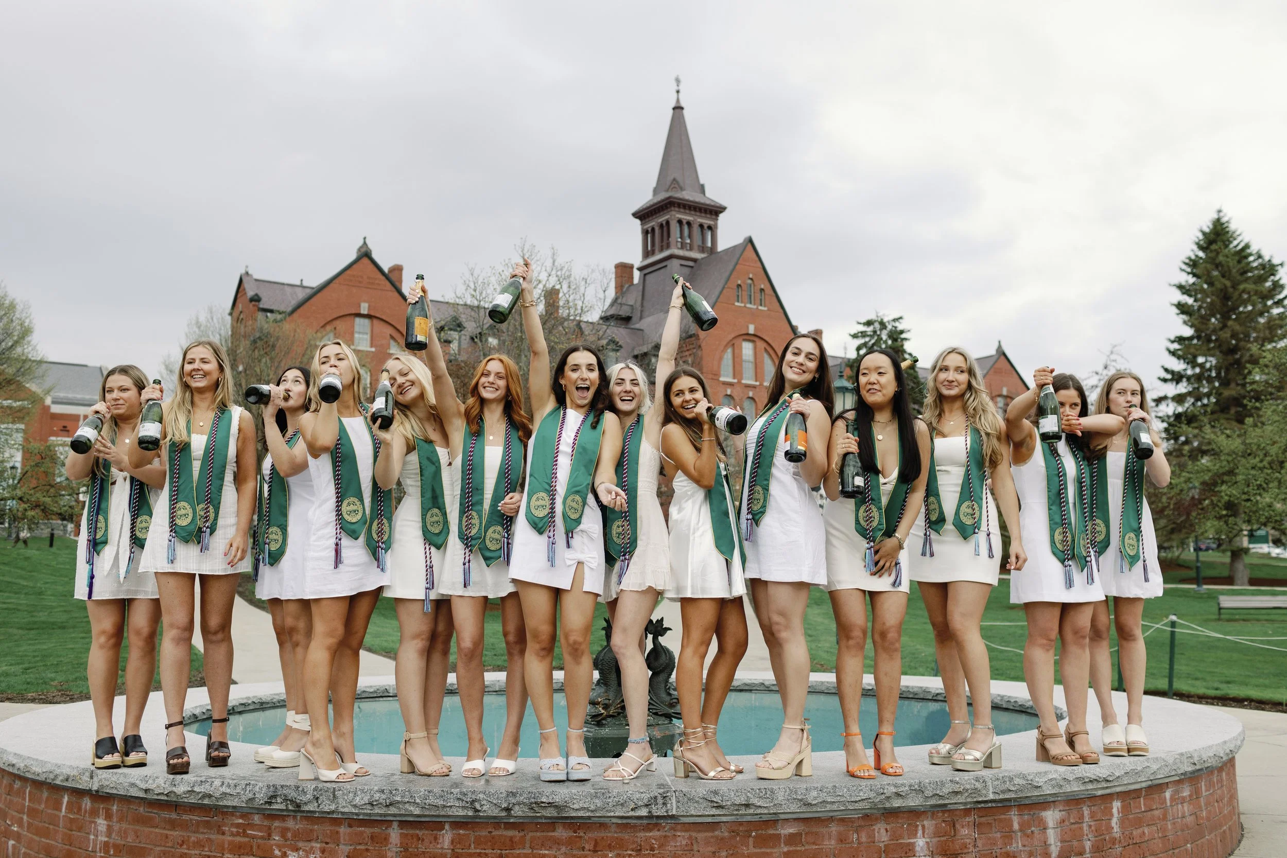 Group of young women in white dresses celebrating graduation, wearing green sashes, holding champagne bottles, standing on a fountain with a historic red brick building in the background, overcast sky.