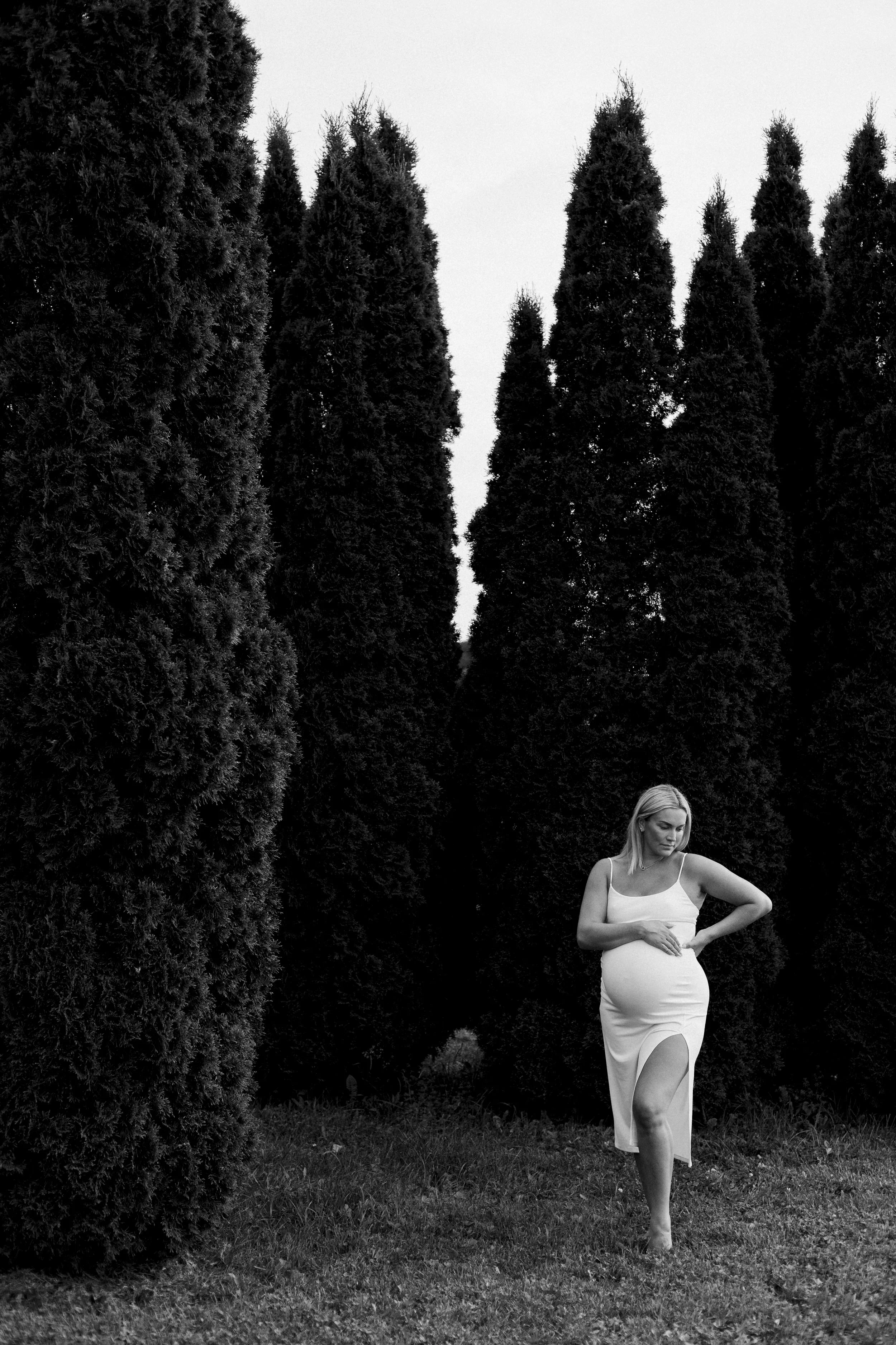 A pregnant woman in a white dress walking barefoot on grass between tall, dark trees.