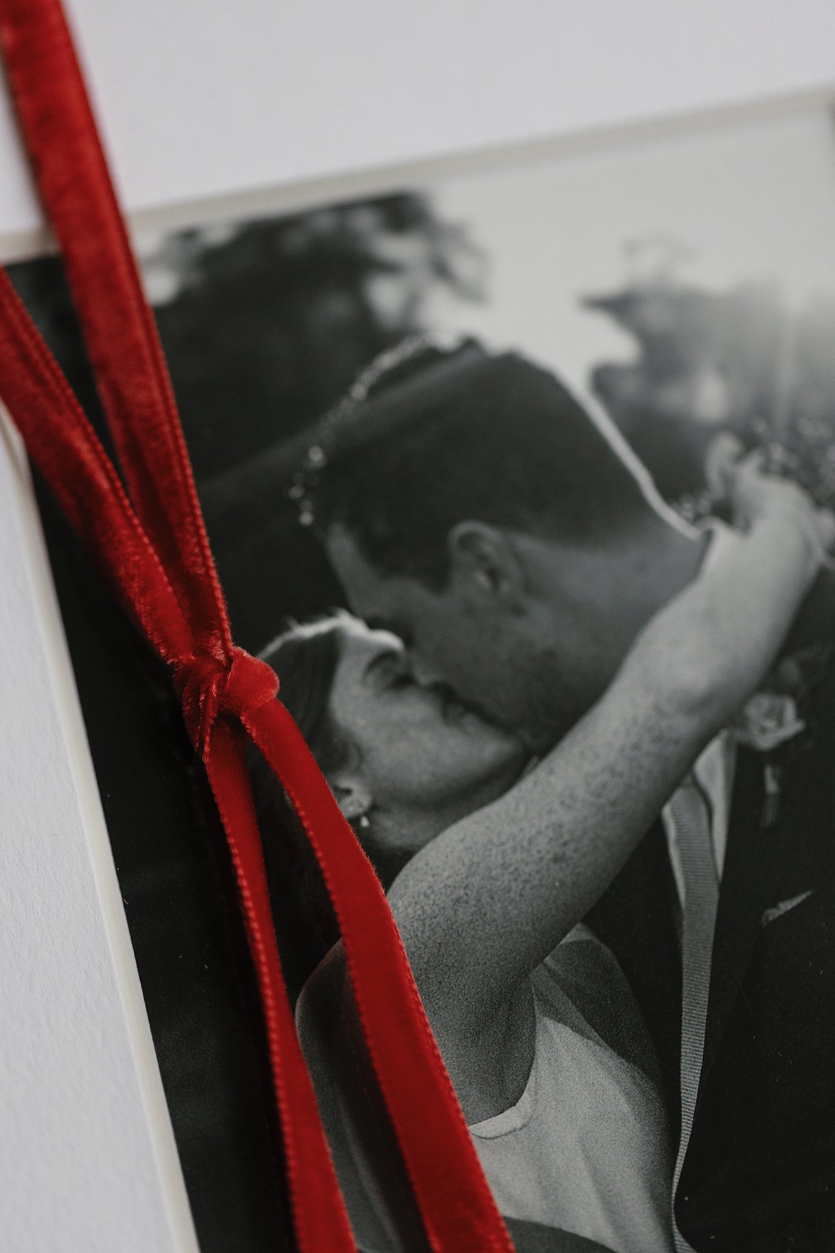 Black and white wedding photo of a couple kissing, with a red ribbon tied around the corner.