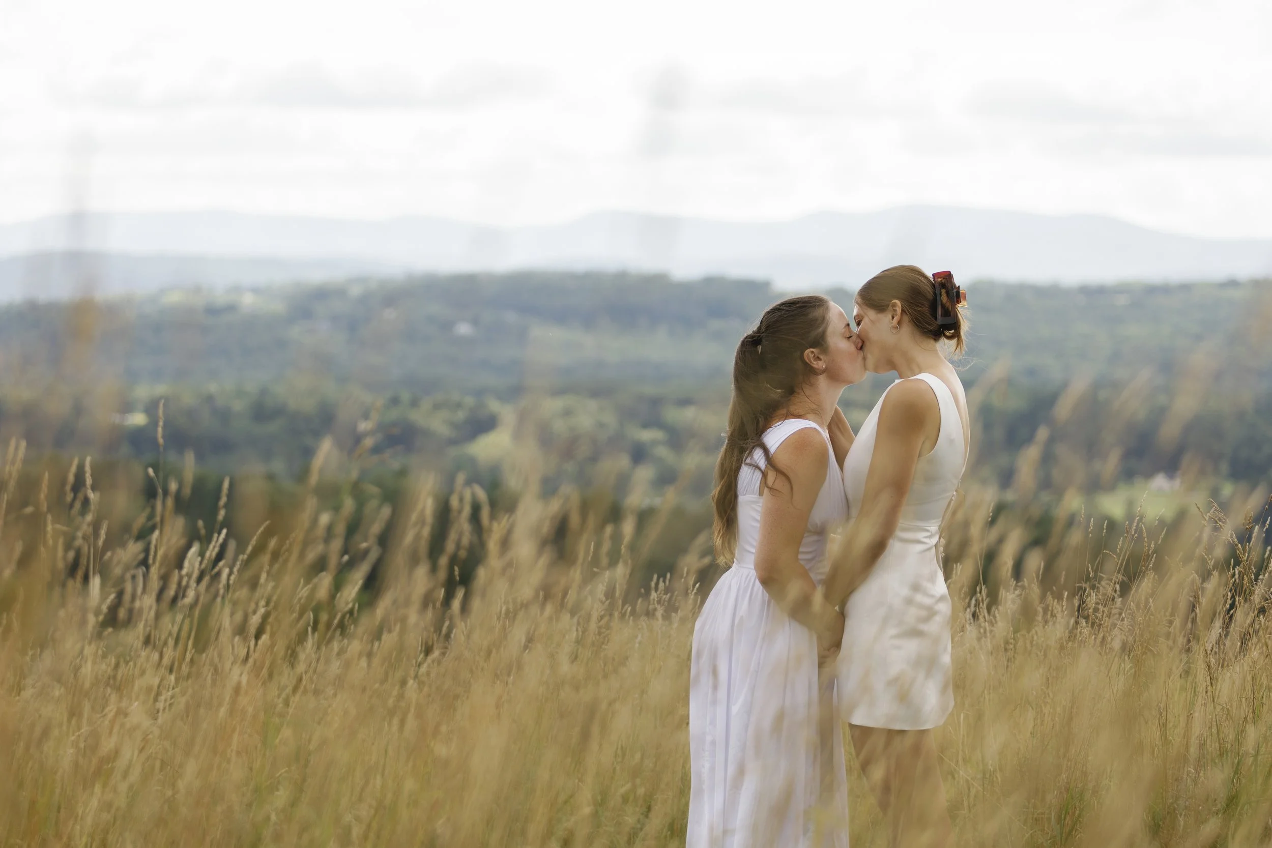 Two women kissing in a field of tall grass with a scenic background of hills and a cloudy sky.