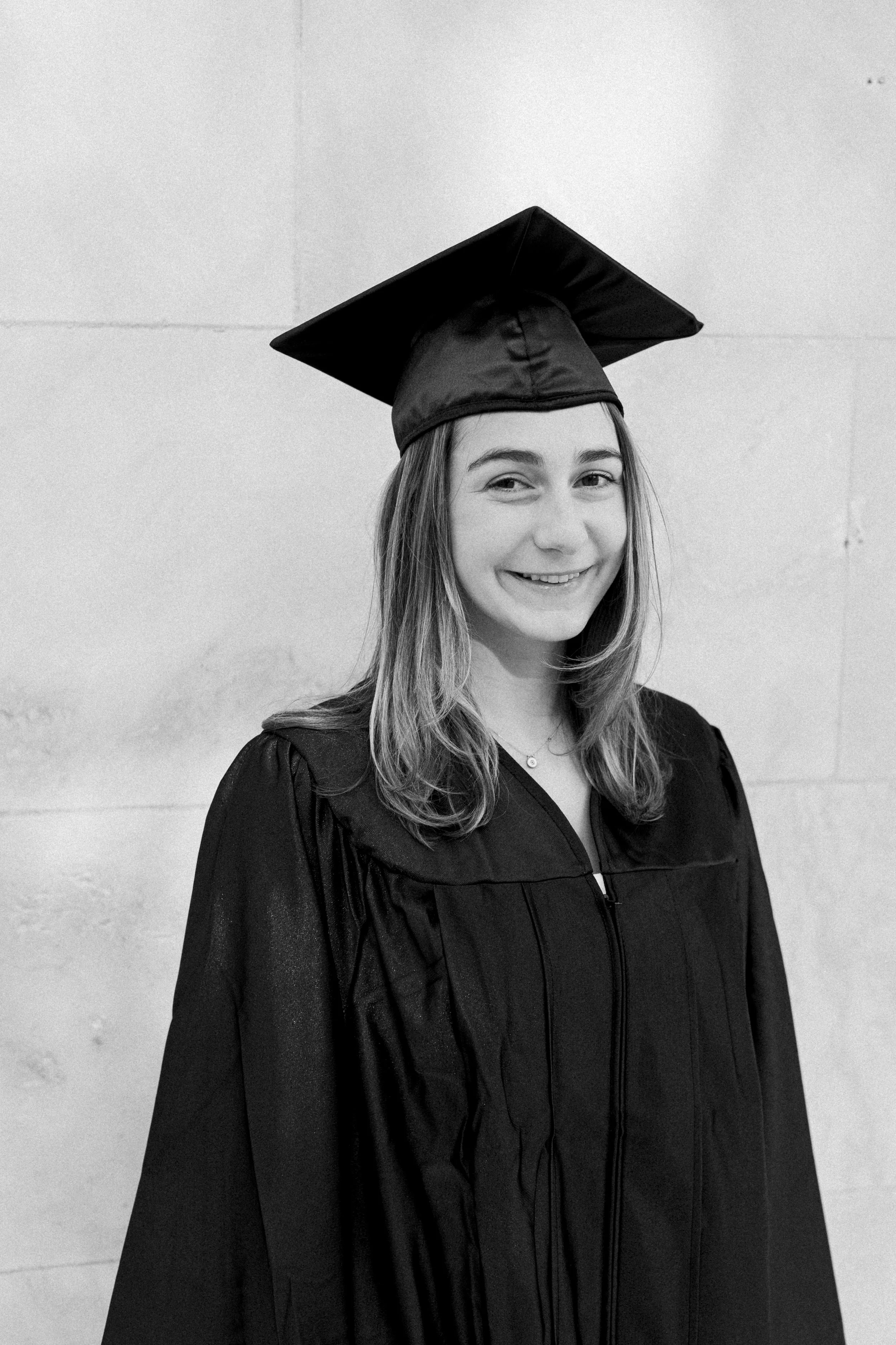 A young woman in graduation cap and gown smiling in front of a plain wall.