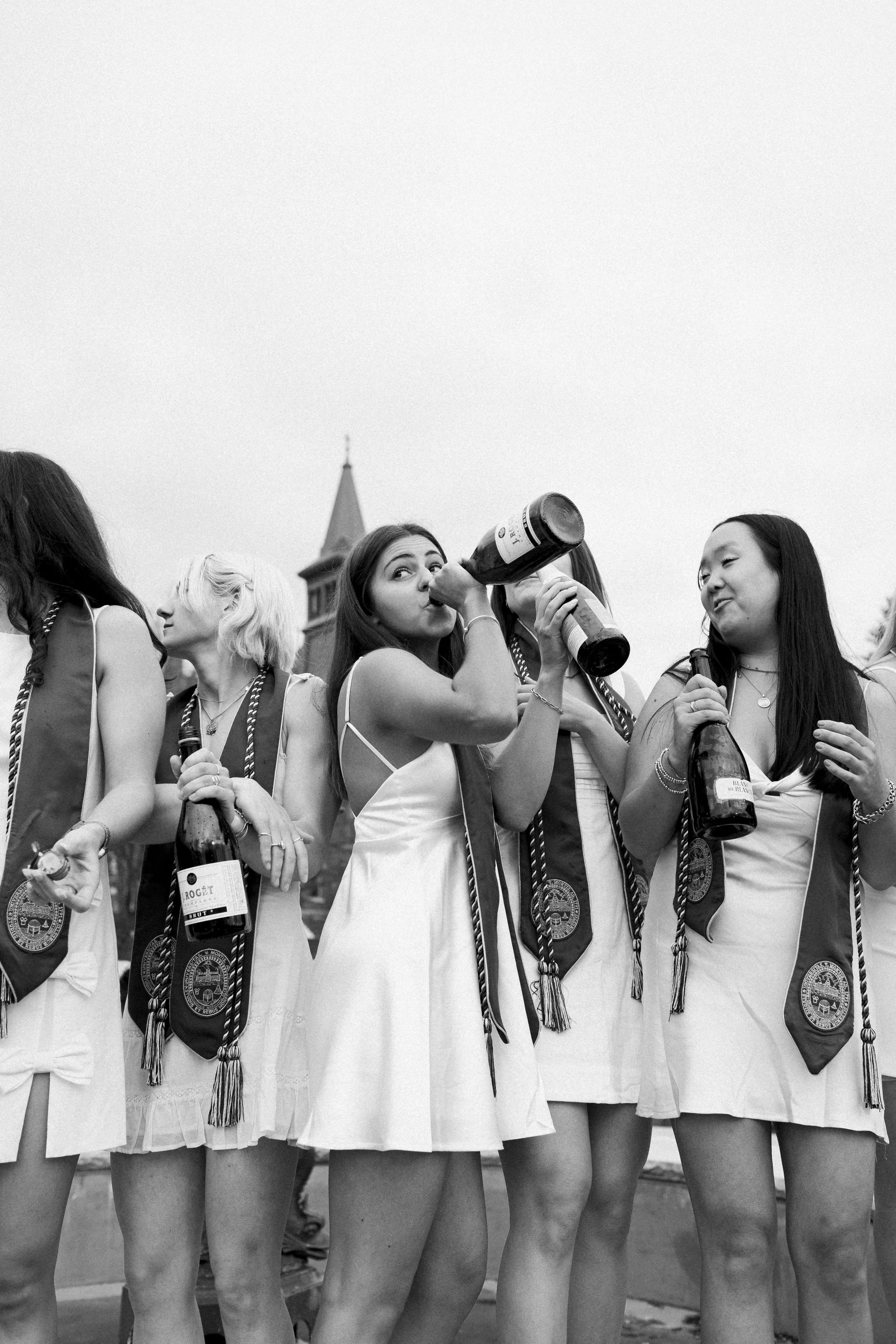 A group of young women in dresses and sashes celebrating at an outdoor event, drinking from bottles, with a university building in the background.