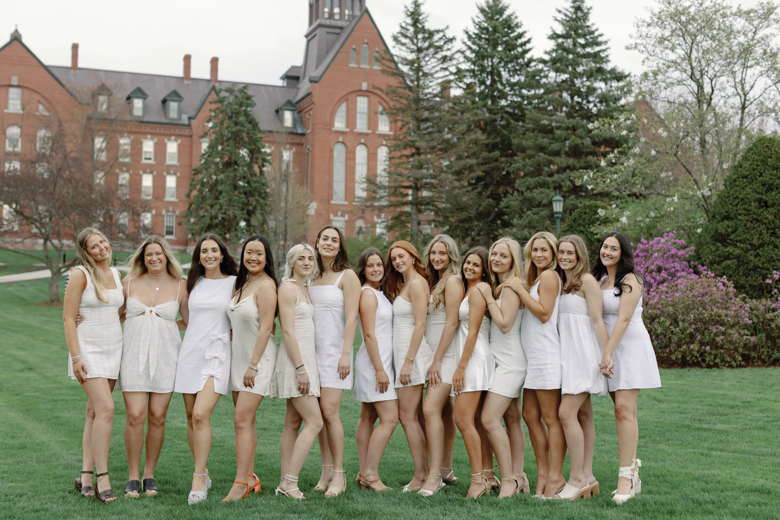 Group of women standing outdoors on a grassy lawn in front of a historic red brick building, dressed in white dresses, smiling and holding hands.