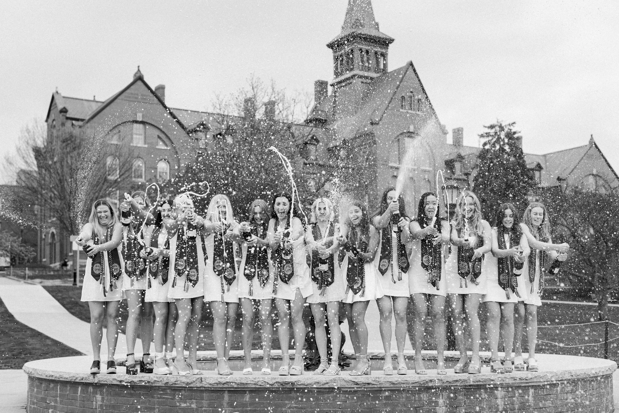 A group of women celebrating on a fountain with champagne bottles, spraying and laughing, in front of a large historic building.
