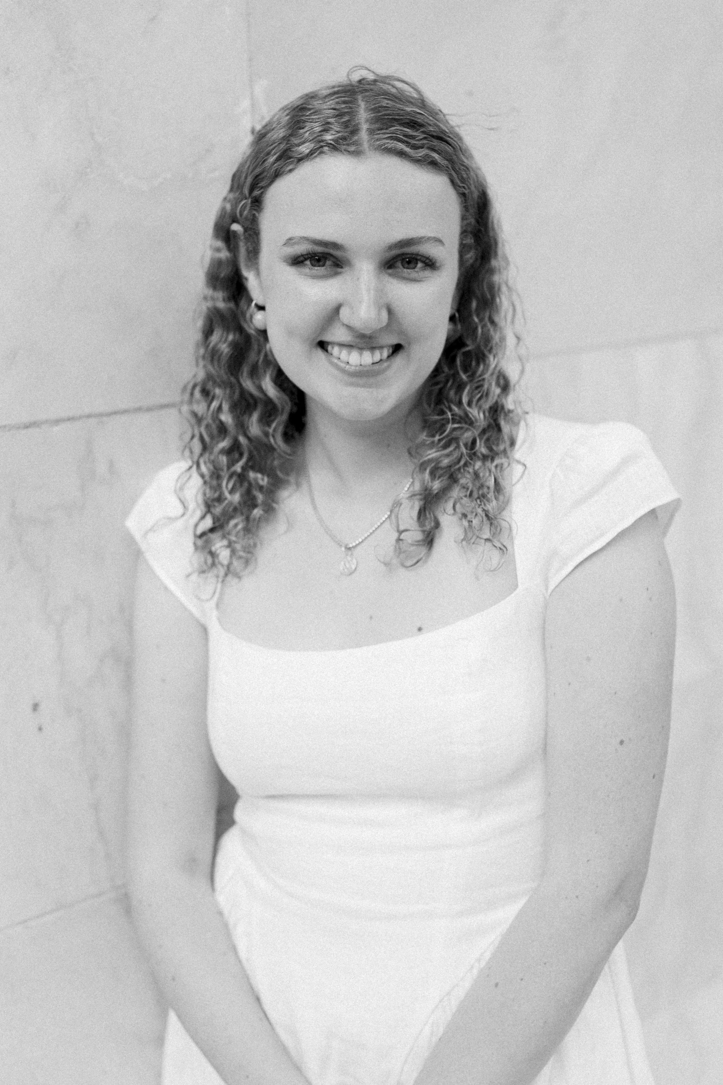 Black and white portrait of a young woman with curly hair, smiling, wearing a white top and a necklace, standing against a plain wall.