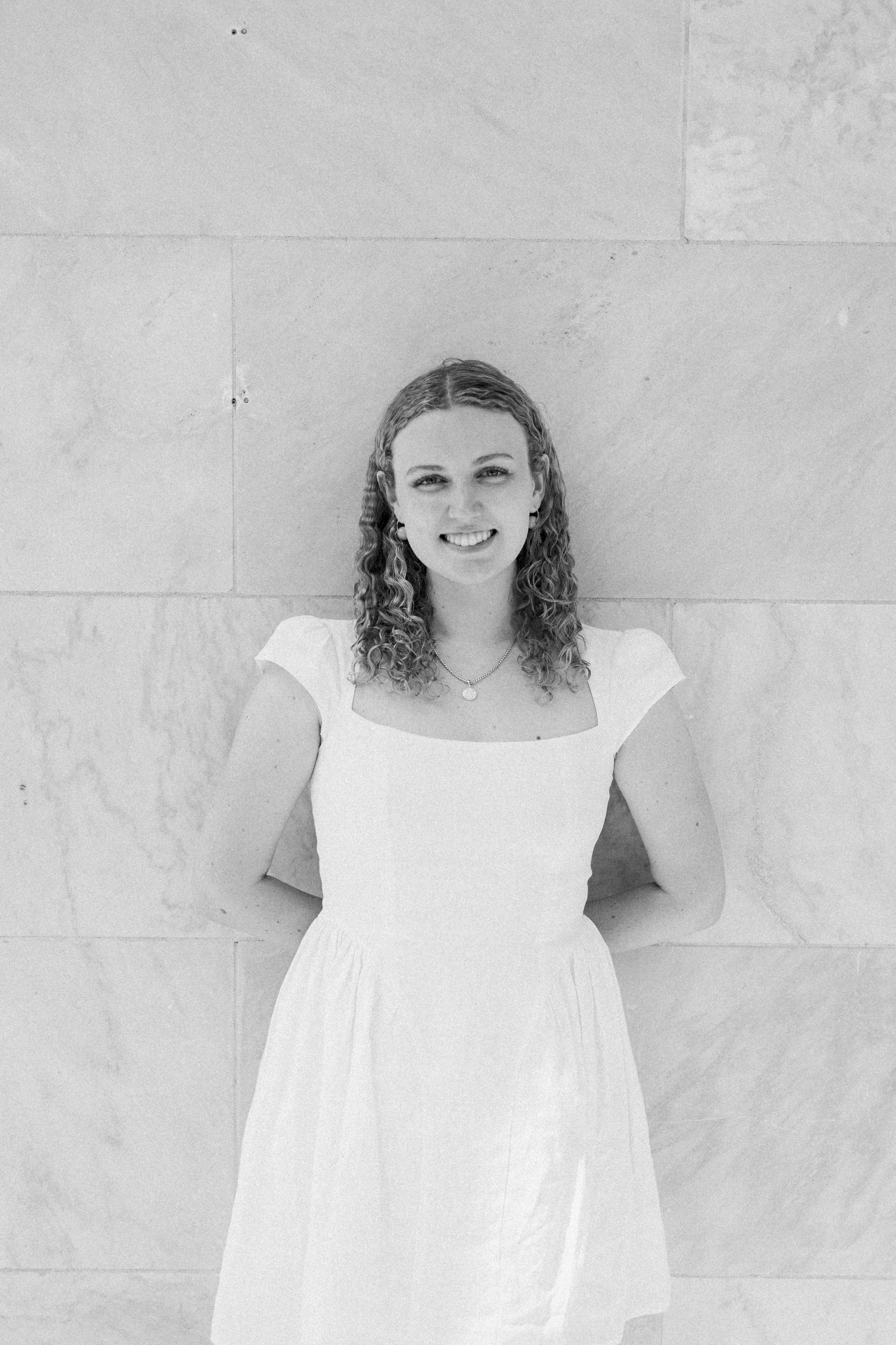 Black and white photo of a young woman with curly hair, smiling, wearing a white dress, standing against a light-colored stone wall with her hands behind her back.