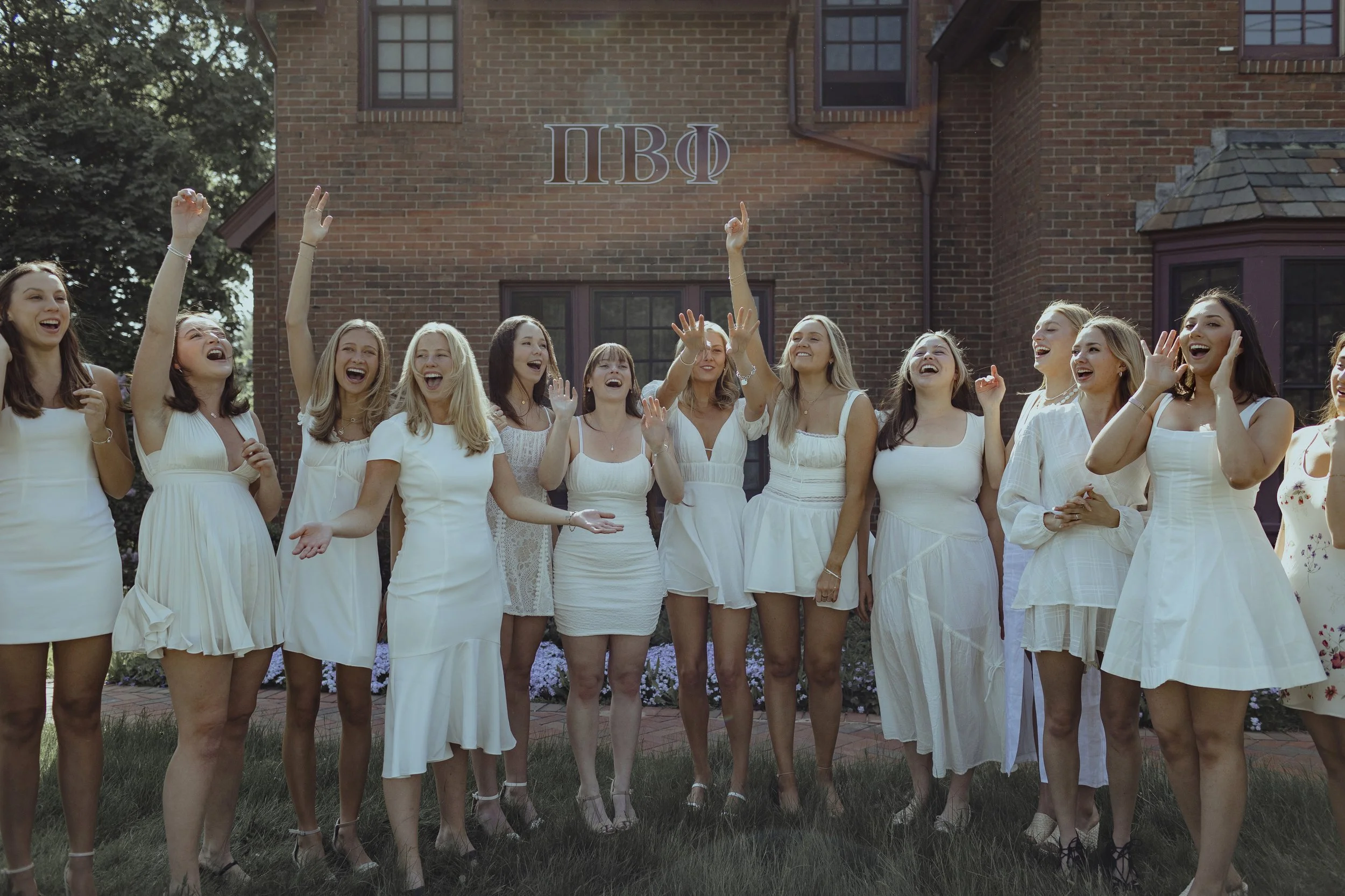 Group of women in white dresses celebrating outdoors in front of a brick house with Greek letters on the wall.