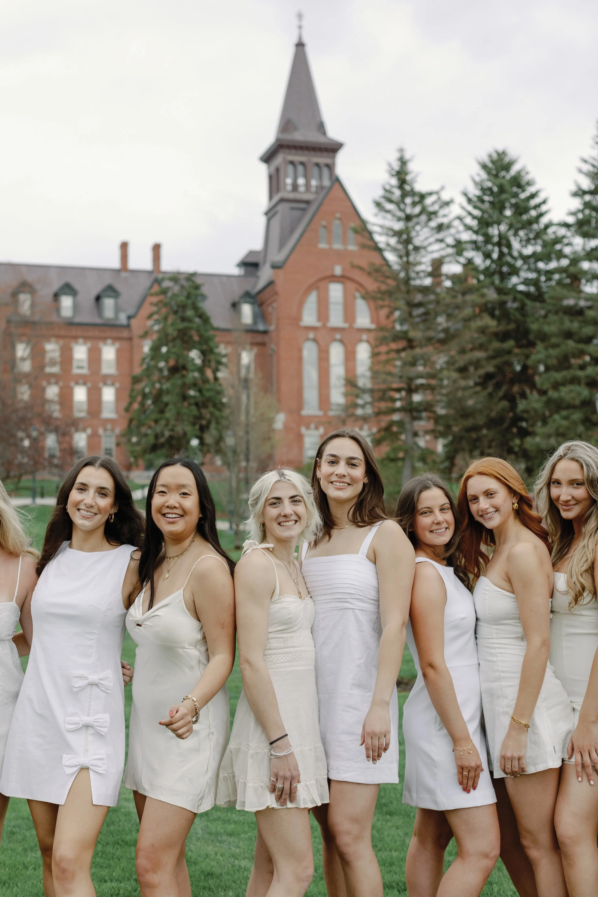 Group of women in white dresses standing on a lush green lawn in front of a large red-brick historic building with a tall spire, trees, and cloudy sky.