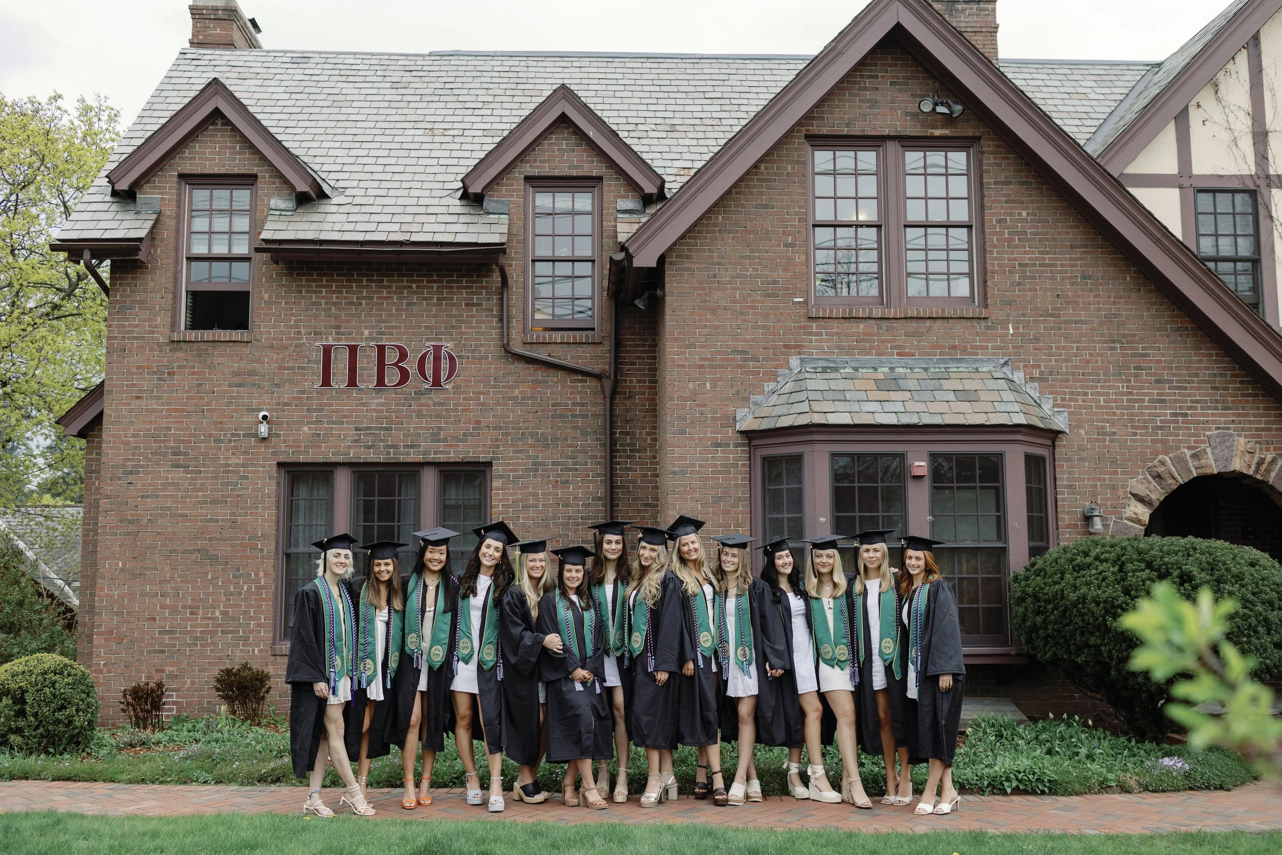 Group of young women in graduation caps and gowns standing in front of a historic brick building with Greek letters 'ΠΒΦ' on the wall.