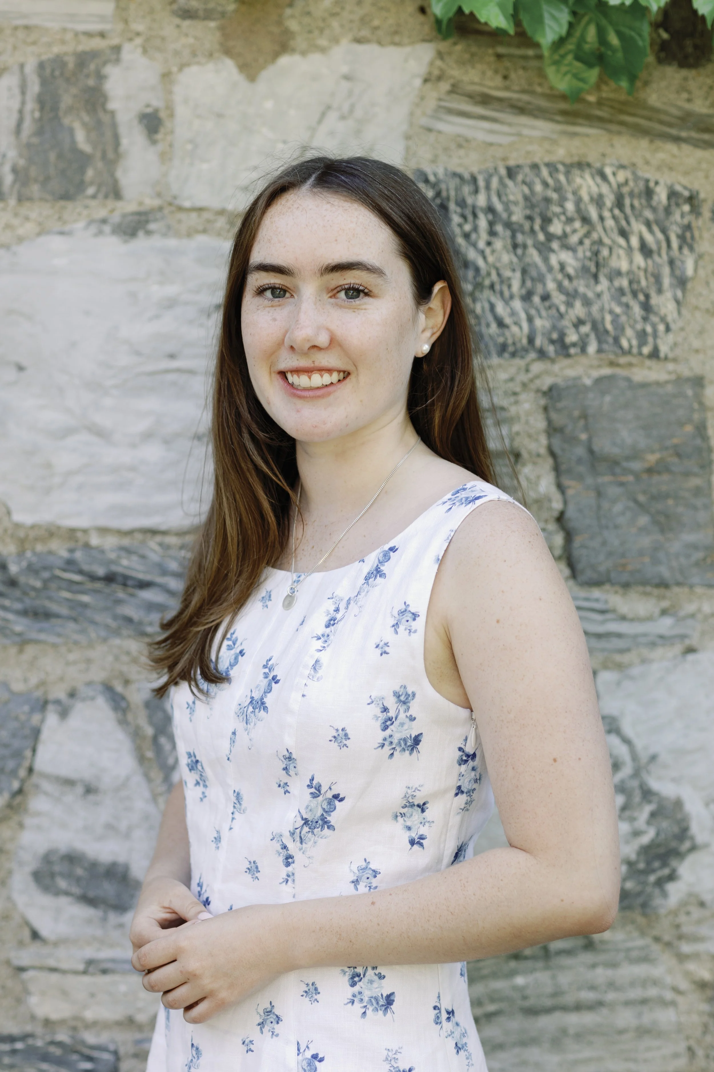 Young woman with long brown hair wearing a white dress with blue floral pattern, standing in front of a stone wall and smiling.