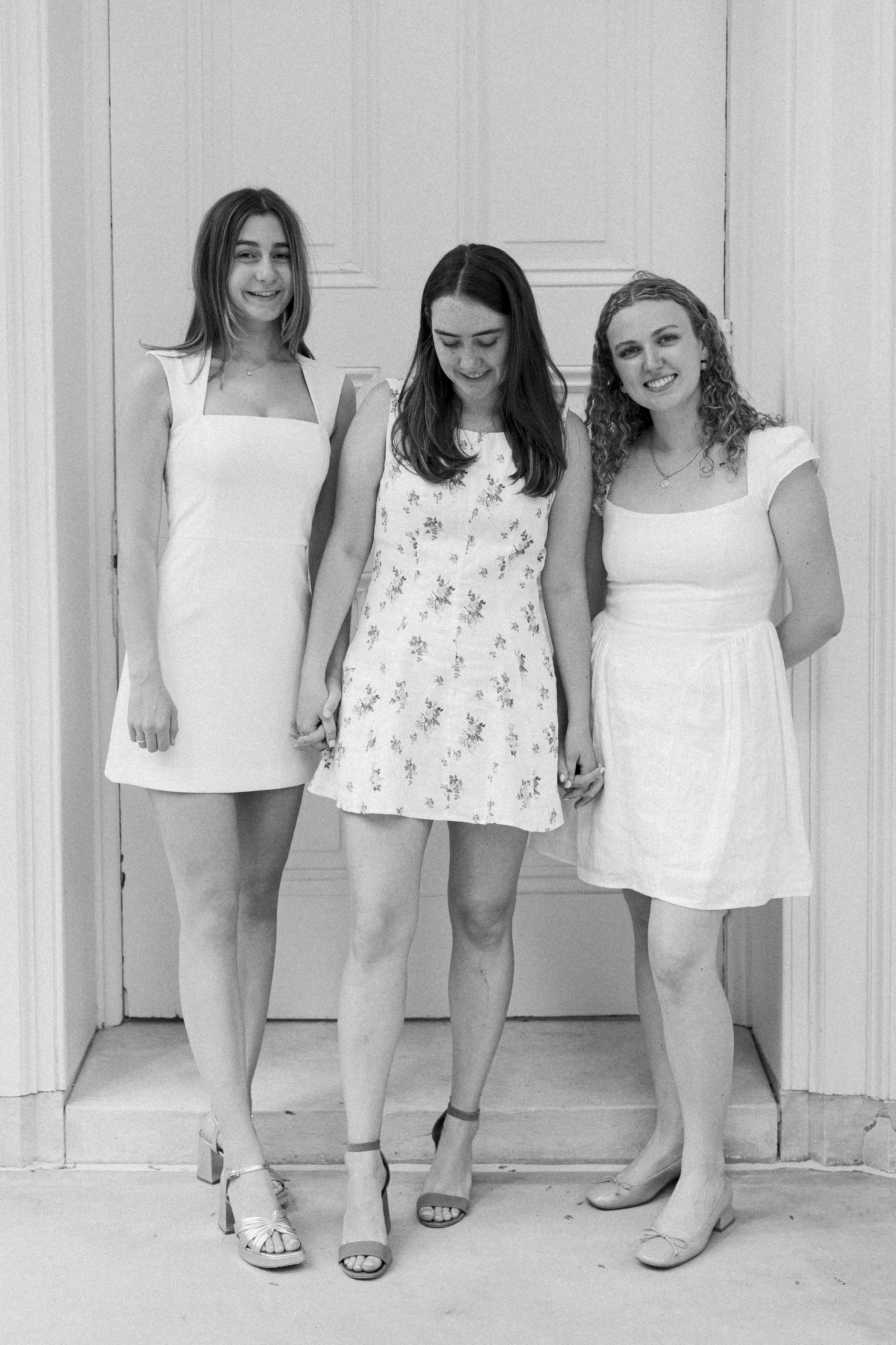 Three young women in dresses standing together in front of a door, holding hands and smiling