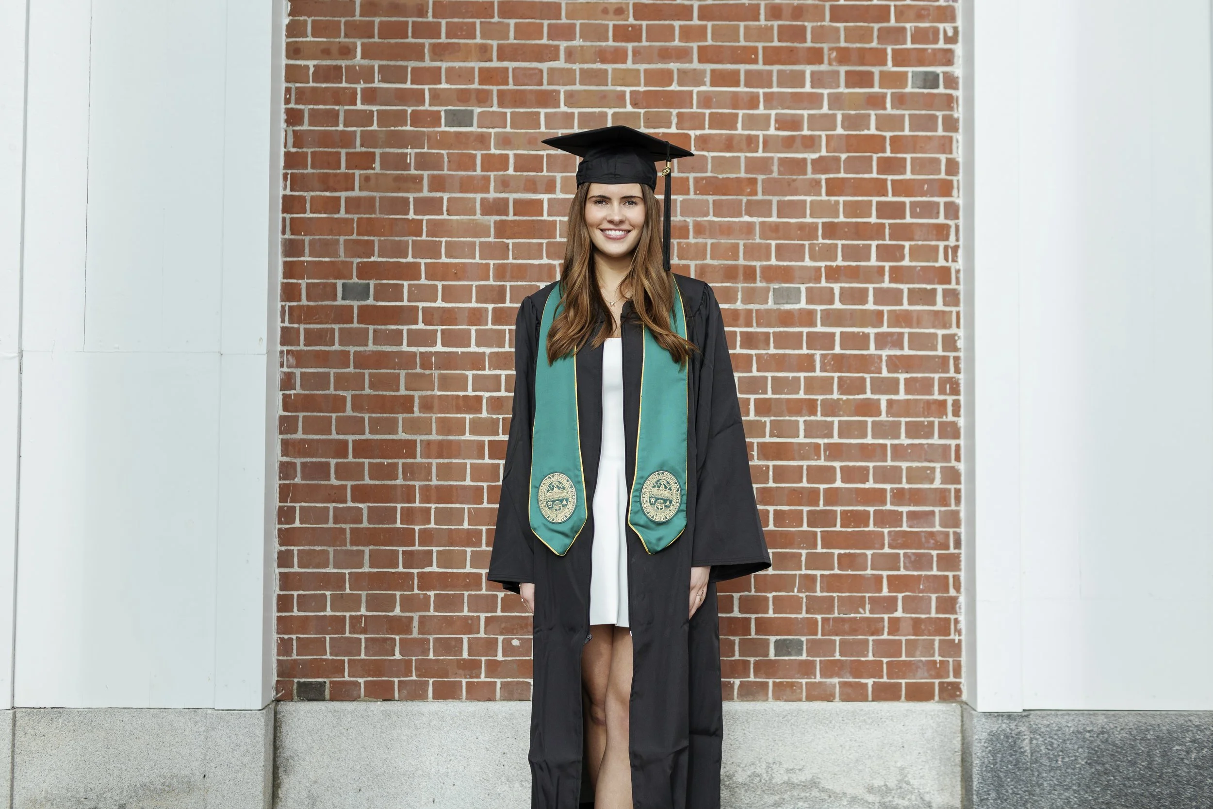 Young woman in cap and gown smiling, standing in front of a brick wall after graduation.