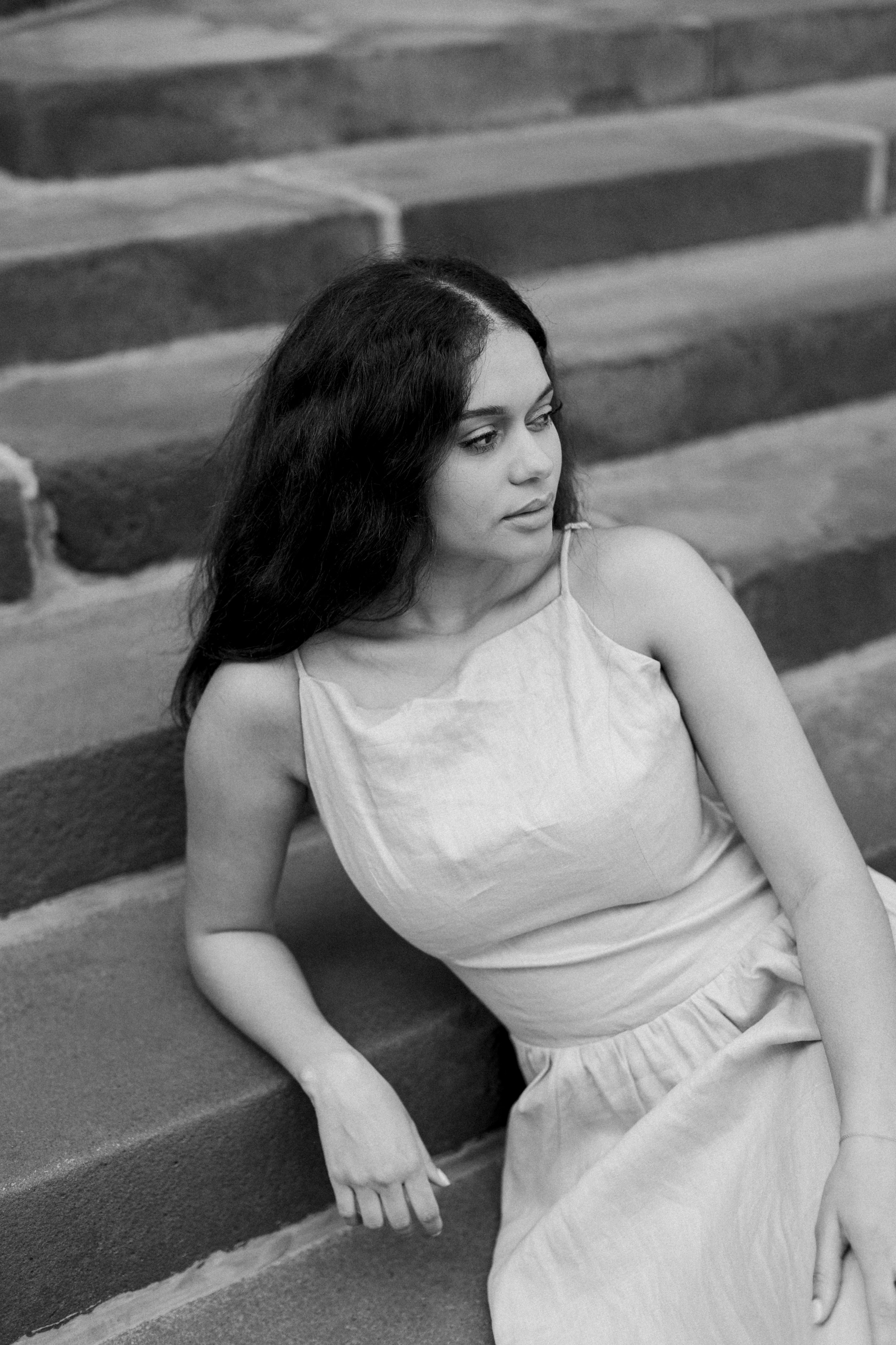 Black and white photo of a young woman with long dark hair, sitting on steps, looking to the side with a calm expression, wearing a sleeveless dress.