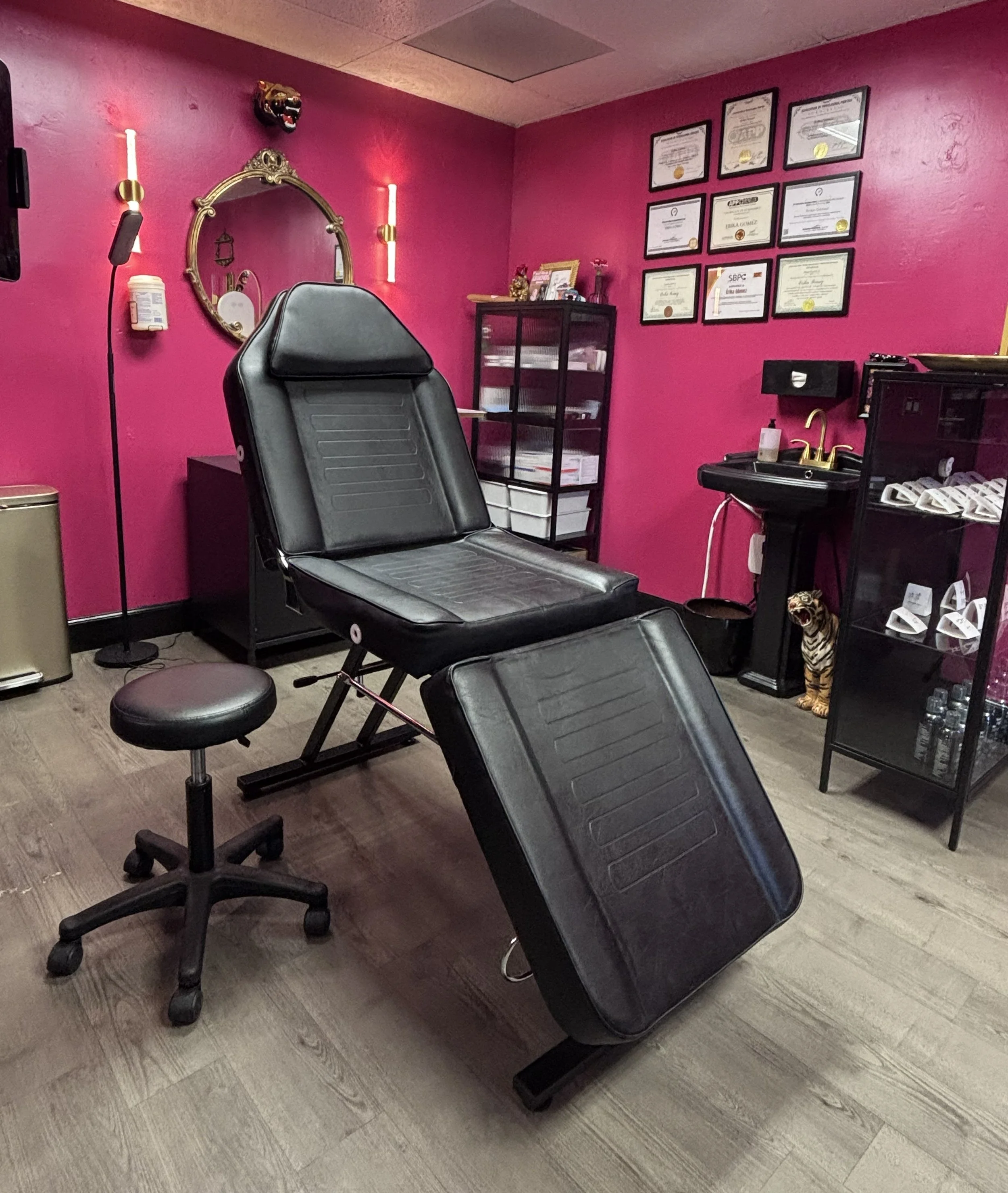 A piercing studio with a black reclining chair, a round stool, a black cabinet with papers, a black counter with a sink and gold faucet, framed certificates on the pink wall, and a tiger statue.
