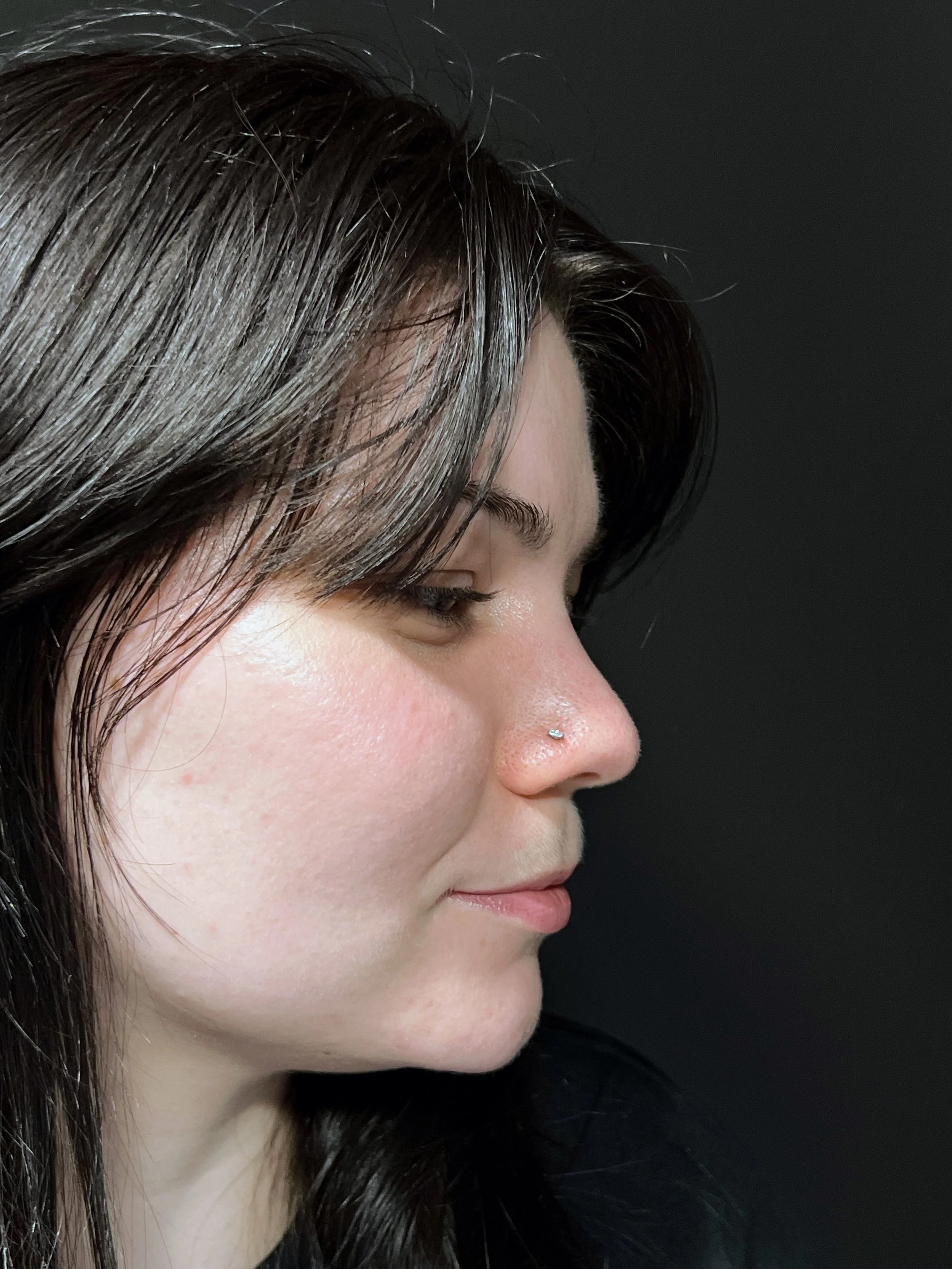 Close-up profile of a woman with dark brown hair and a nose piercing, against a black background.