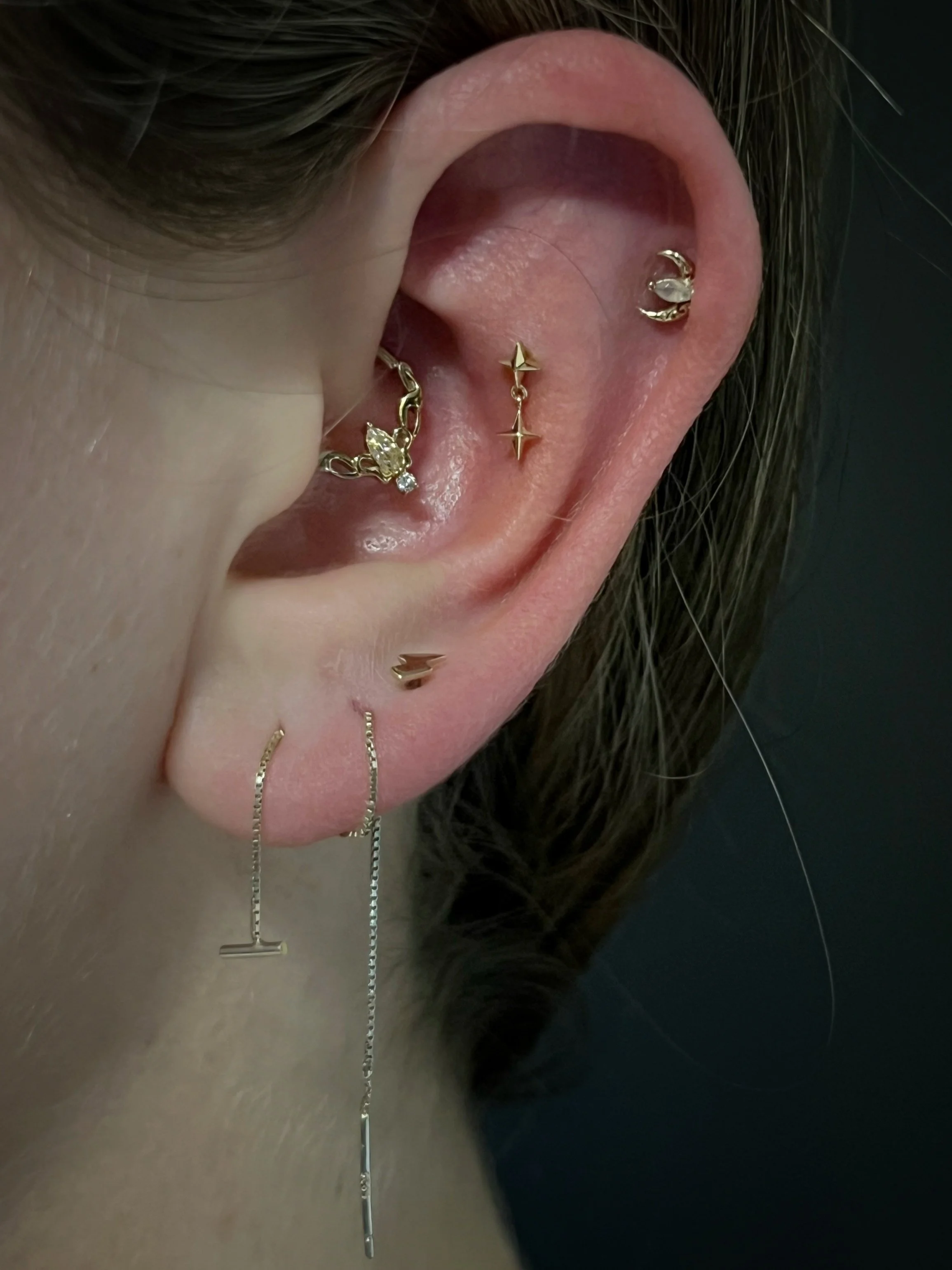 Close-up of a person's ear with multiple earrings, including a star-shaped earring, a crescent moon earring, and a flower-shaped earring with a gemstone. Several thin chains are dangling from some earrings.