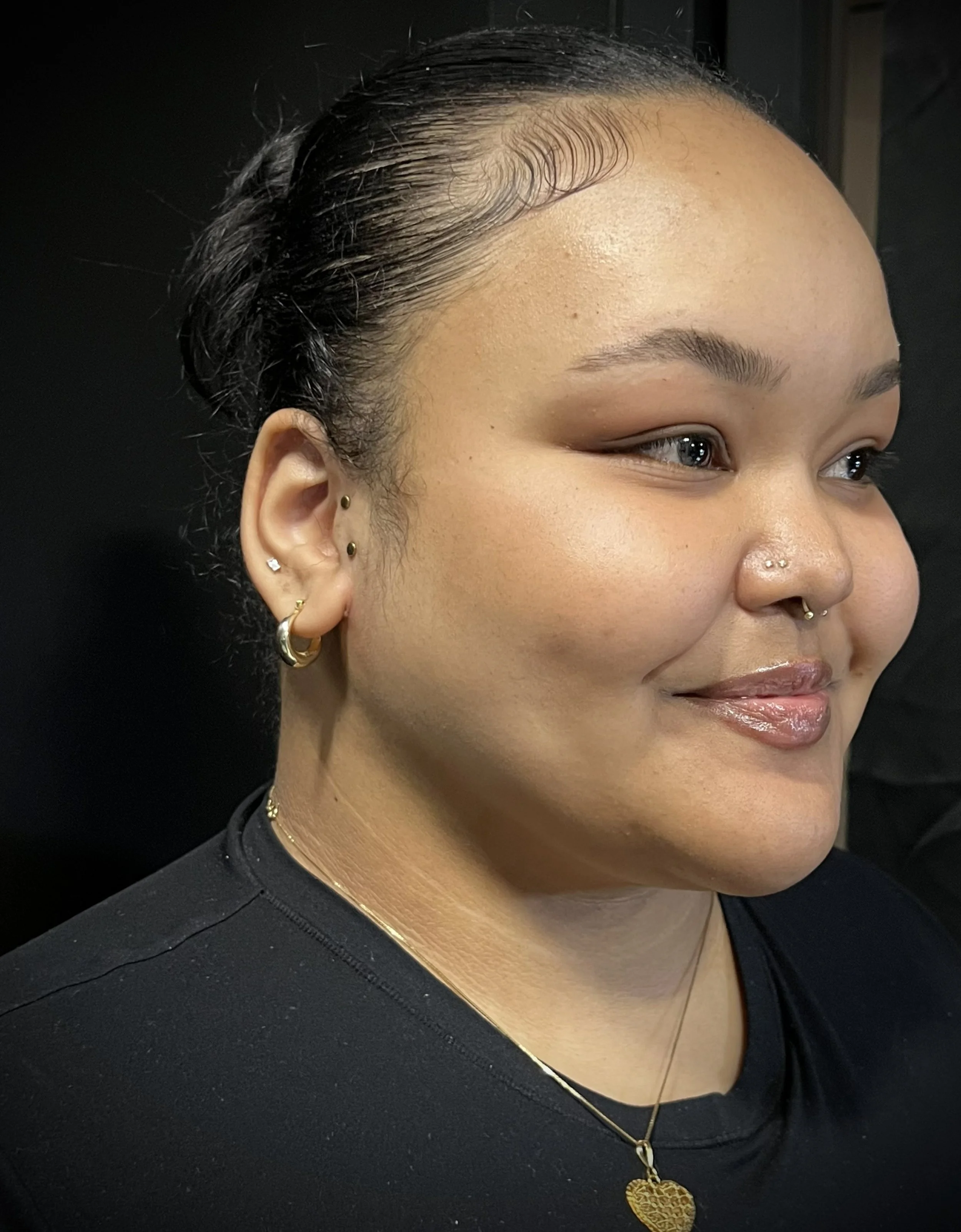 Close-up of a woman's face showing her right side, with black hair slicked back, multiple earrings in her ear, a nose piercing, and a gold heart-shaped necklace, against a dark background.