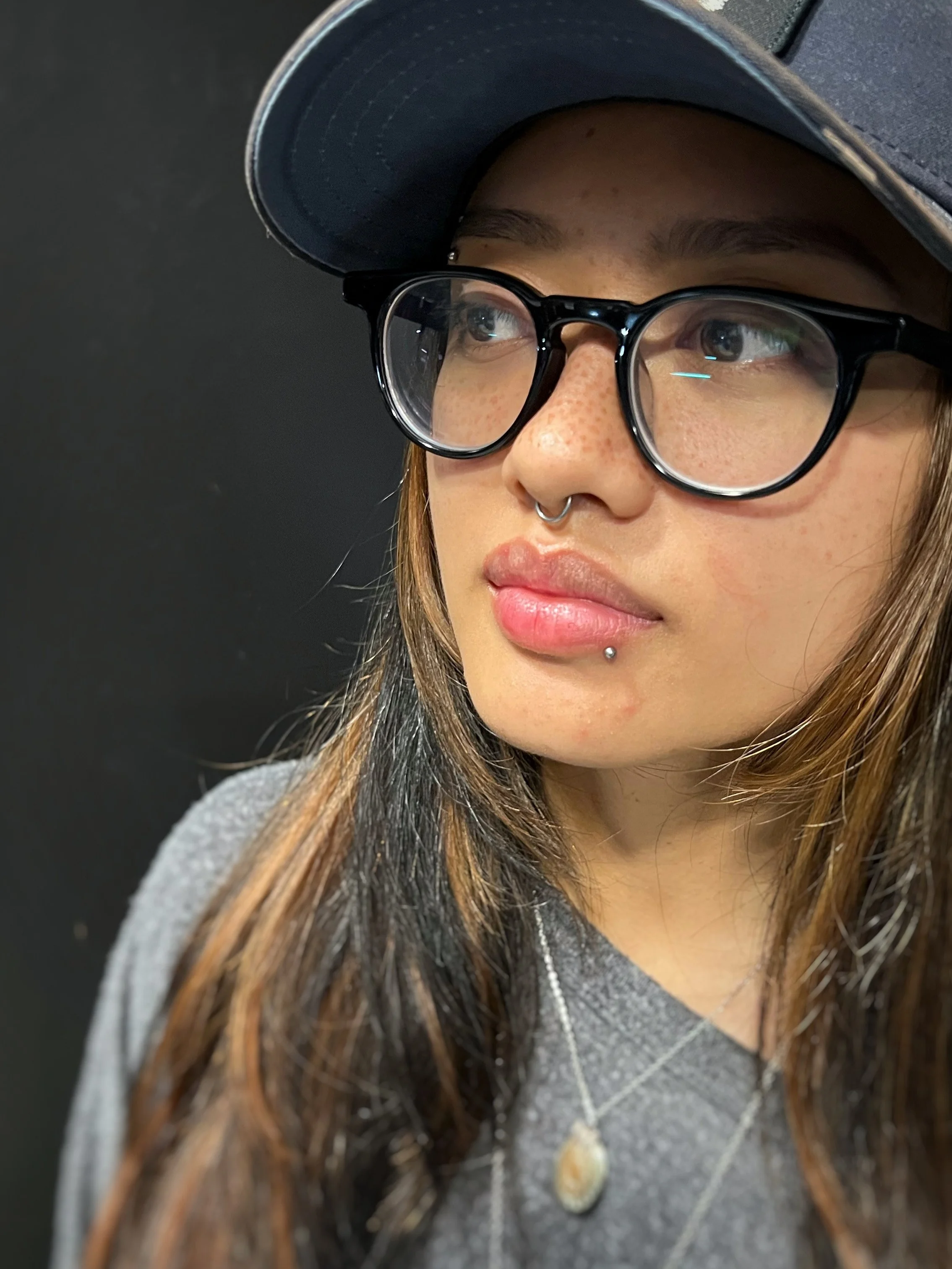 A close-up of a young woman wearing black glasses and a cap, with long brown hair, freckles, and piercings, looking to her right against a dark background.