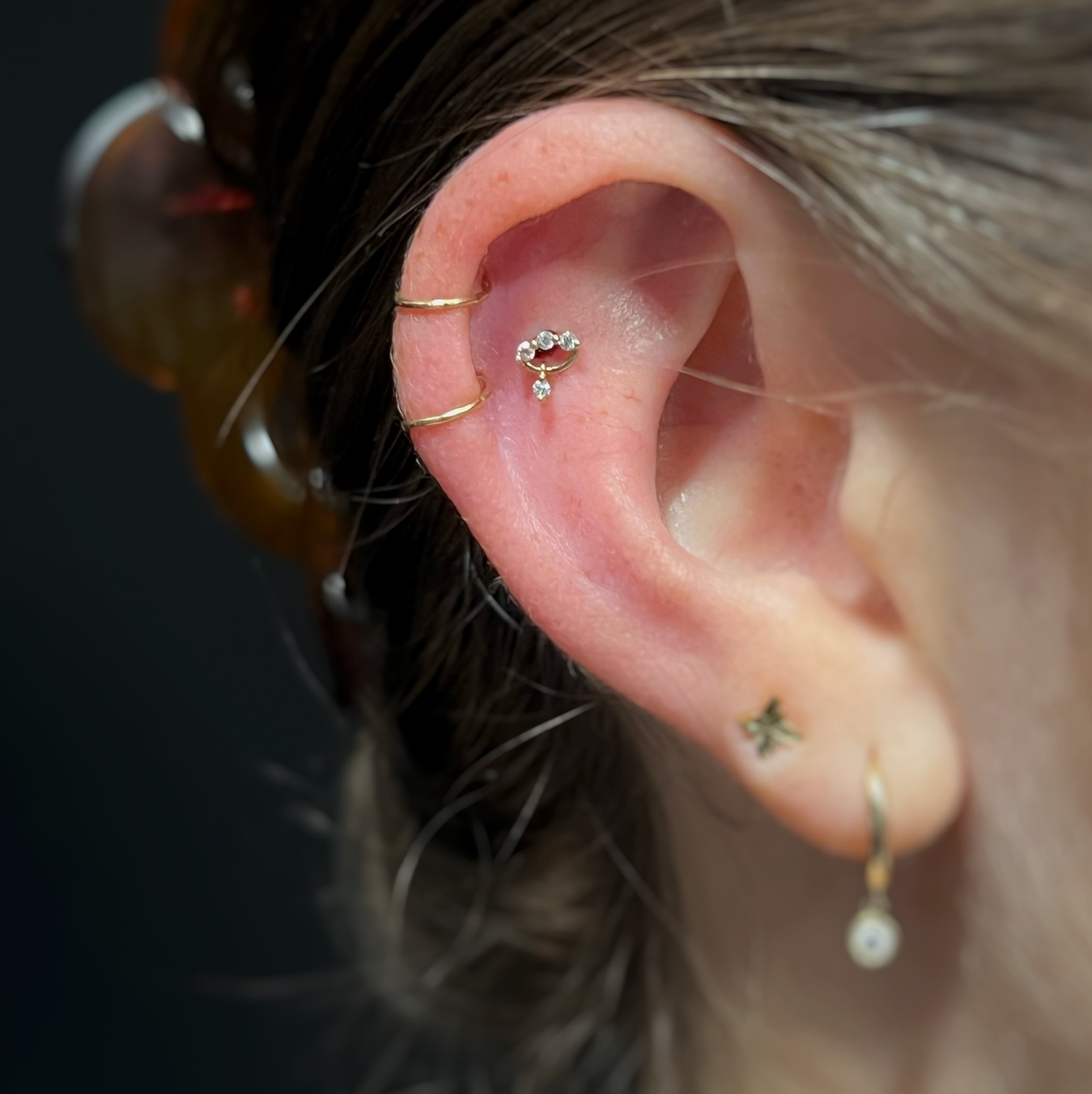 Close-up of a person's ear with a small diamond hoop earring and a cartilage piercing adorned with a tiny silver ring. The person has dark hair with highlighted streaks, and there are hairpins visible. The background is blurred.
