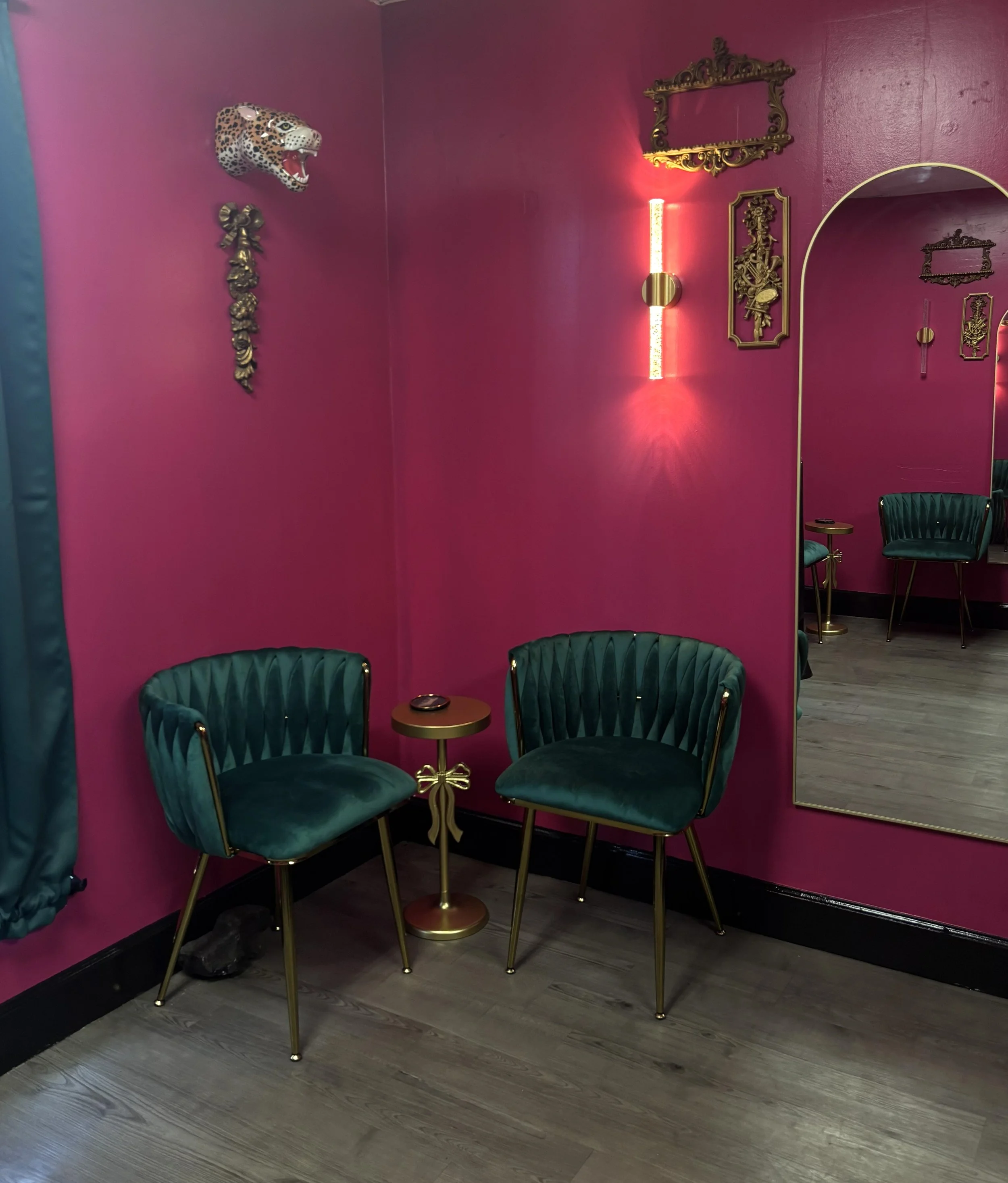 A pink wall accented with gold ornate decor, including a jaguar head wall mount, a vertical light fixture, and framed artwork, with teal chairs and a small gold table in front of a mirror reflecting the same decor.