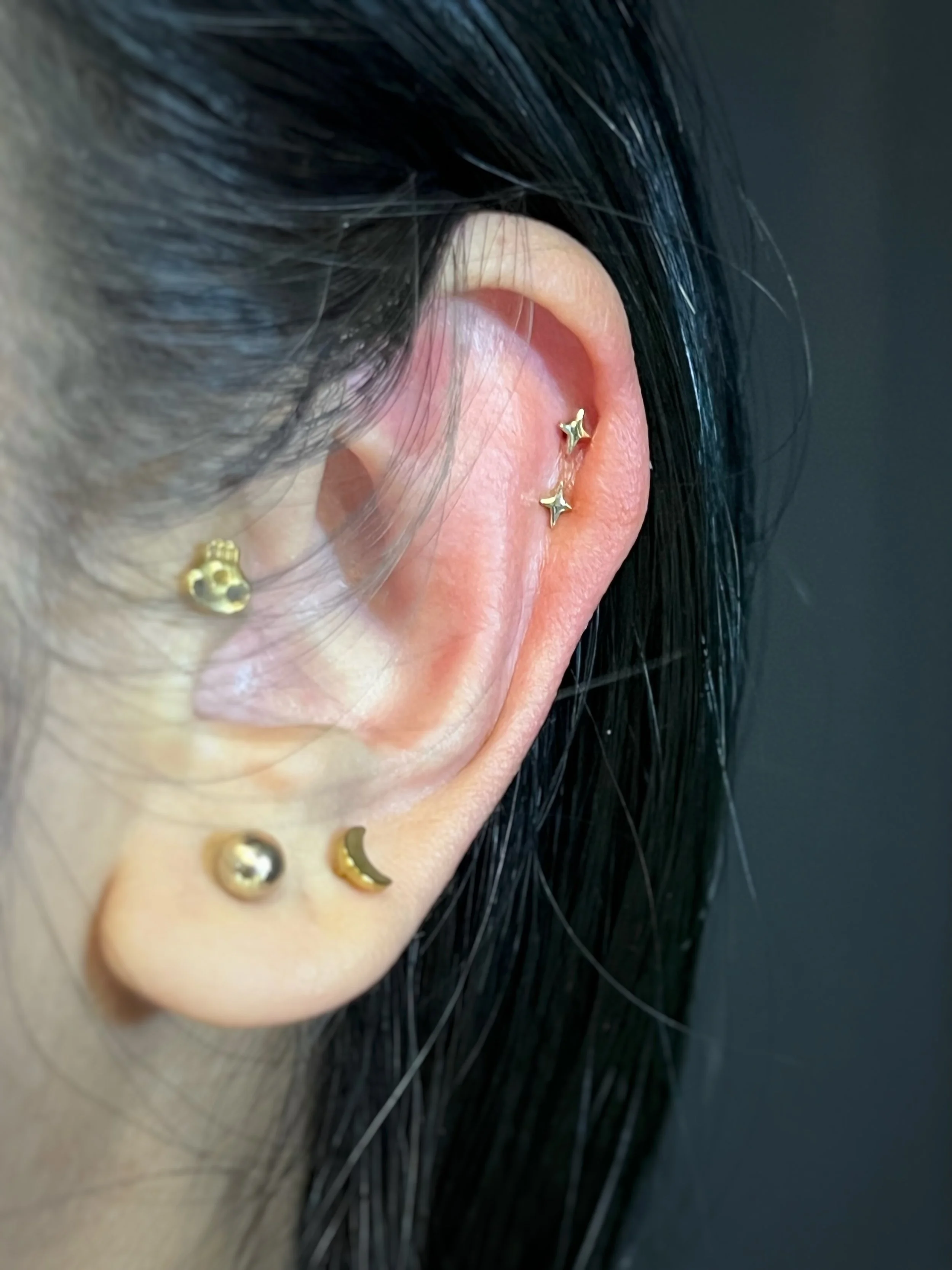 Close-up image of a person's ear with a gold star-shaped cartilage piercing and other small earrings, alongside dark hair.