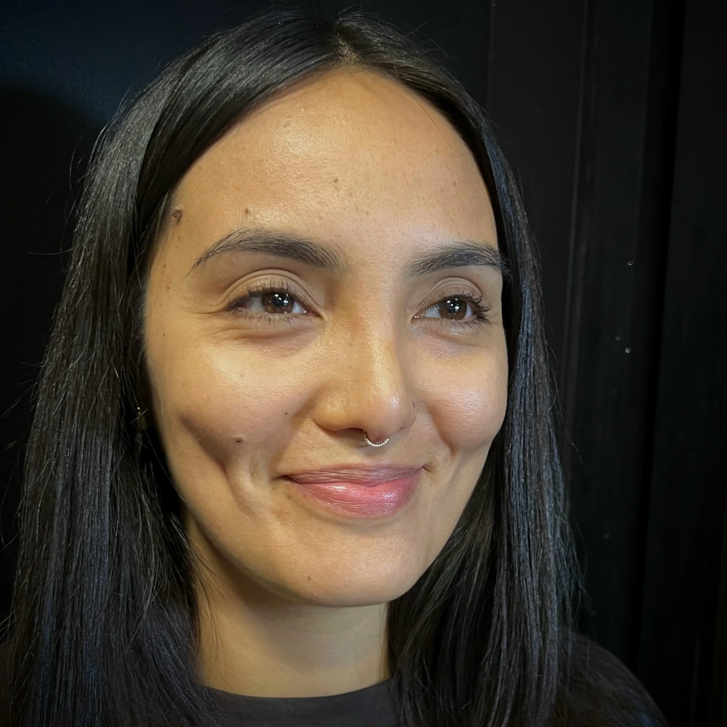 Close-up of a woman's face with dark hair, brown eyes, light makeup, and a septum piercing, against a black background.