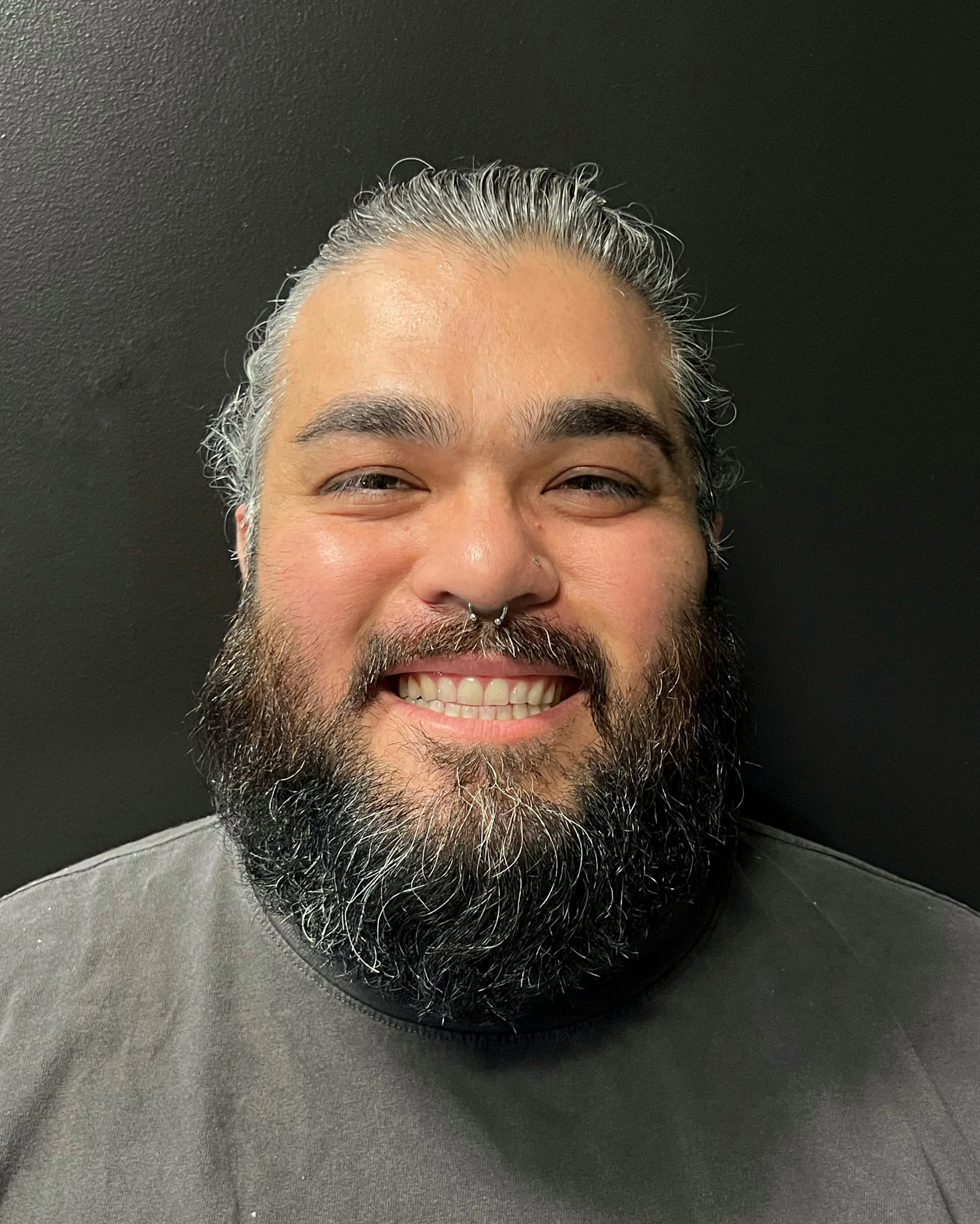 A smiling man with a septum piercing, with gray and black hair and a full beard, wearing a dark shirt, standing against a black background.
