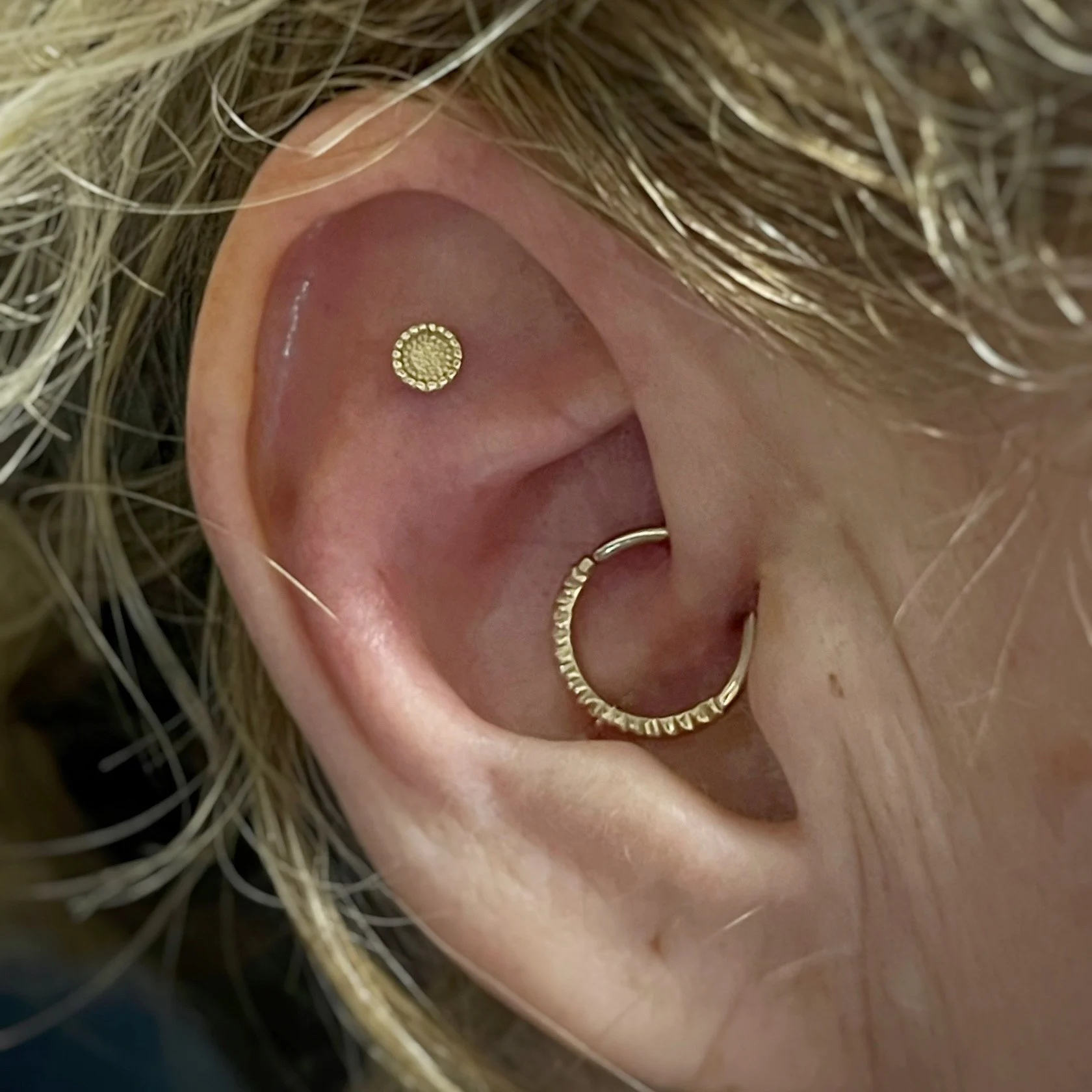 Close-up of a person's ear with a gold hoop earring and a stud piercing, surrounded by light-colored hair.